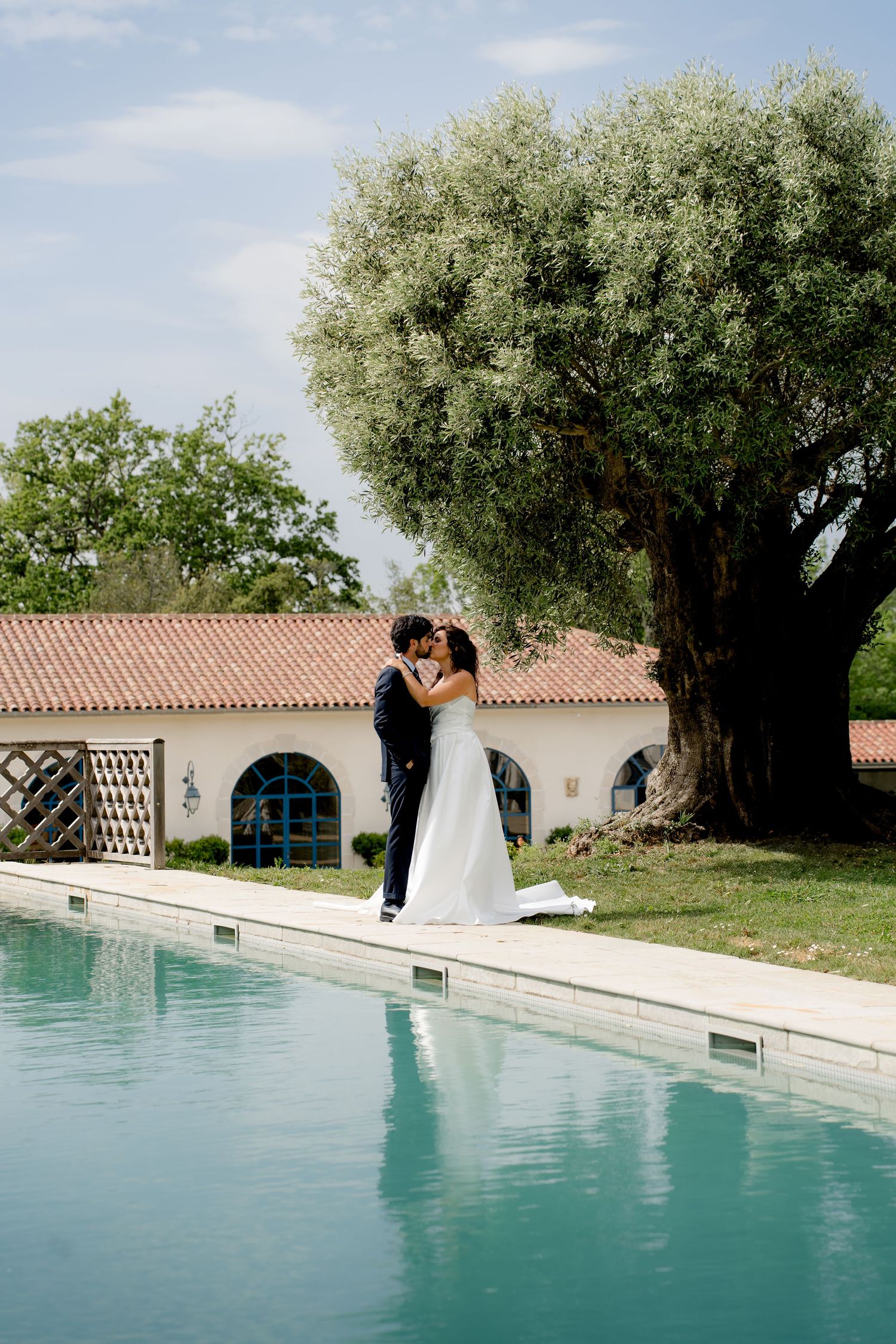 Newlyweds share a tender kiss beside a sparkling pool under a large olive tree at a rustic Italian villa.