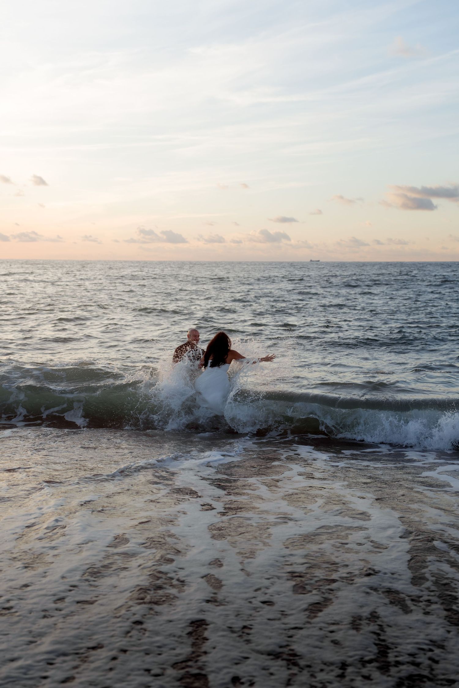 Couple splashing playfully in ocean waves at dusk during romantic beach photo shoot.