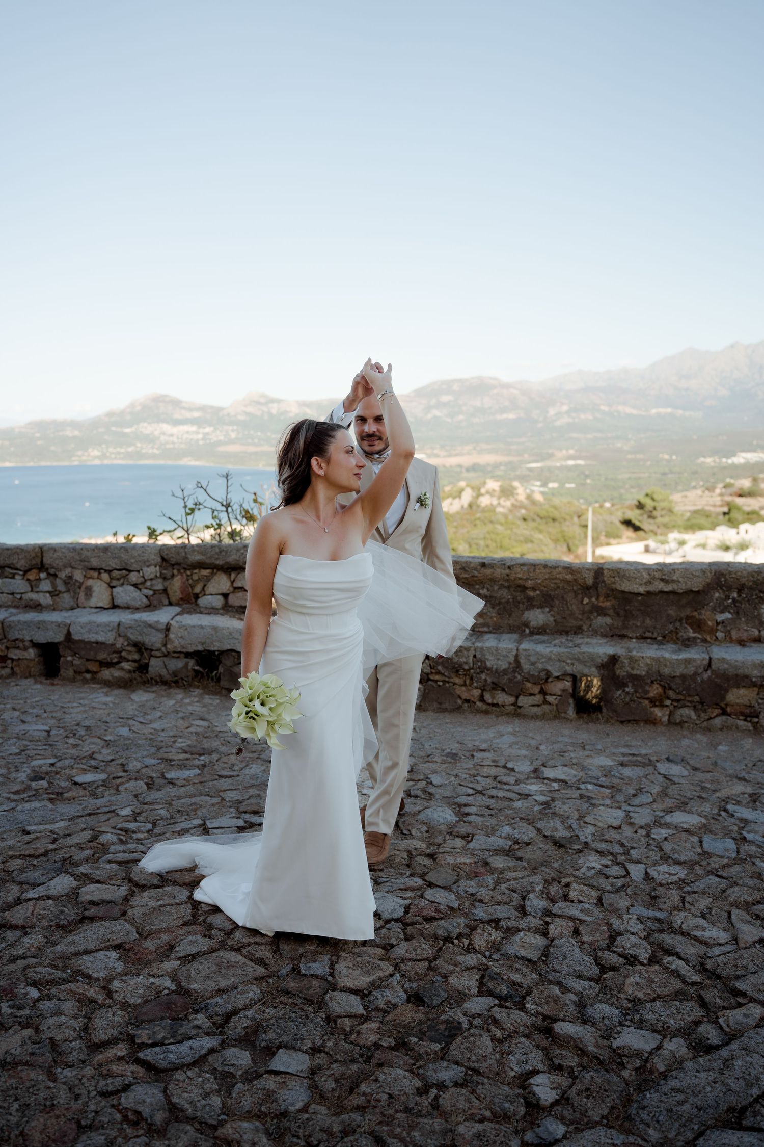 Groom spinning bride in white dress on rustic stone terrace with mountain and sea views.