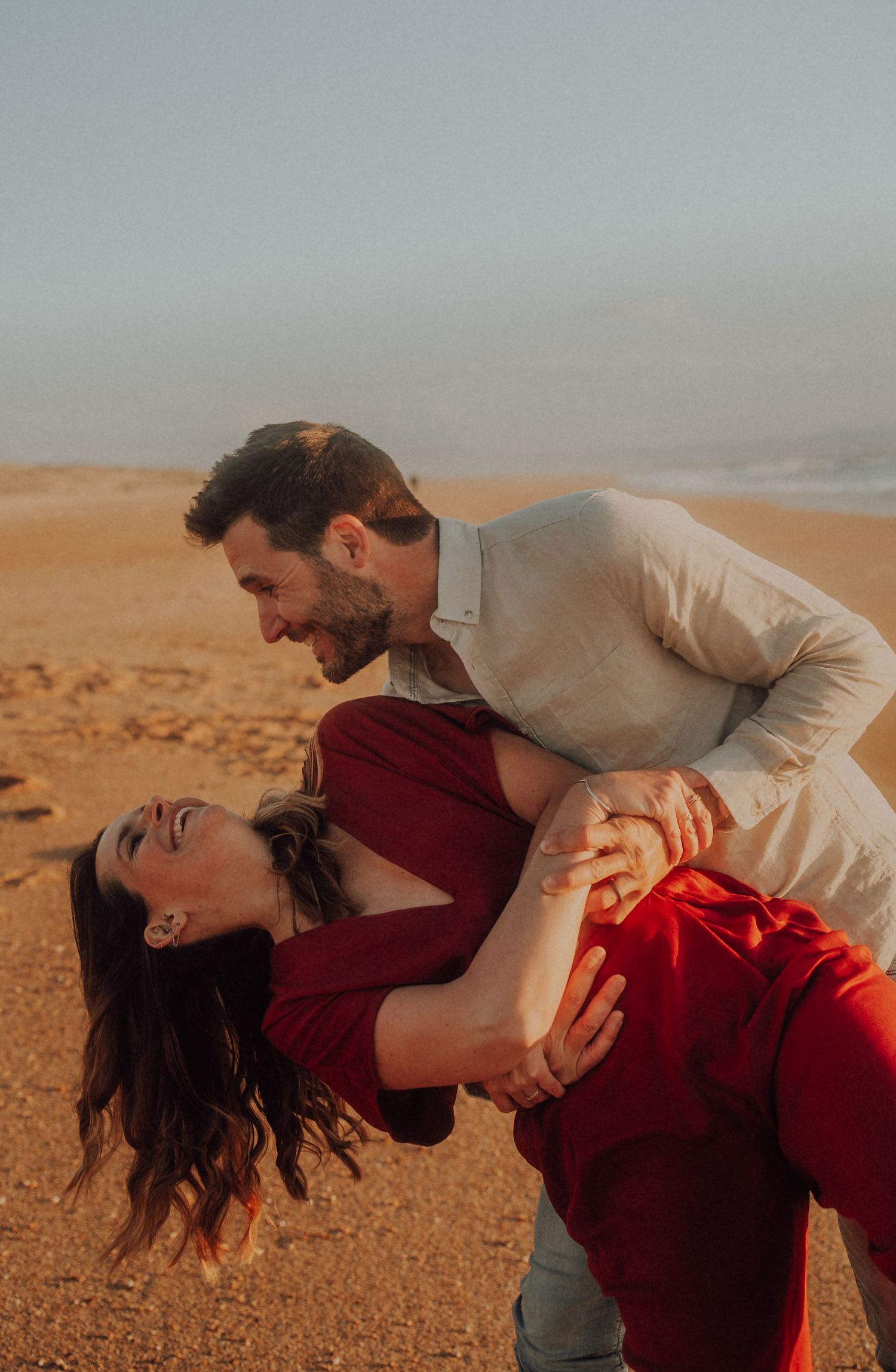 Couple laughing and dancing on a sandy beach at sunset, man dipping woman in red dress.