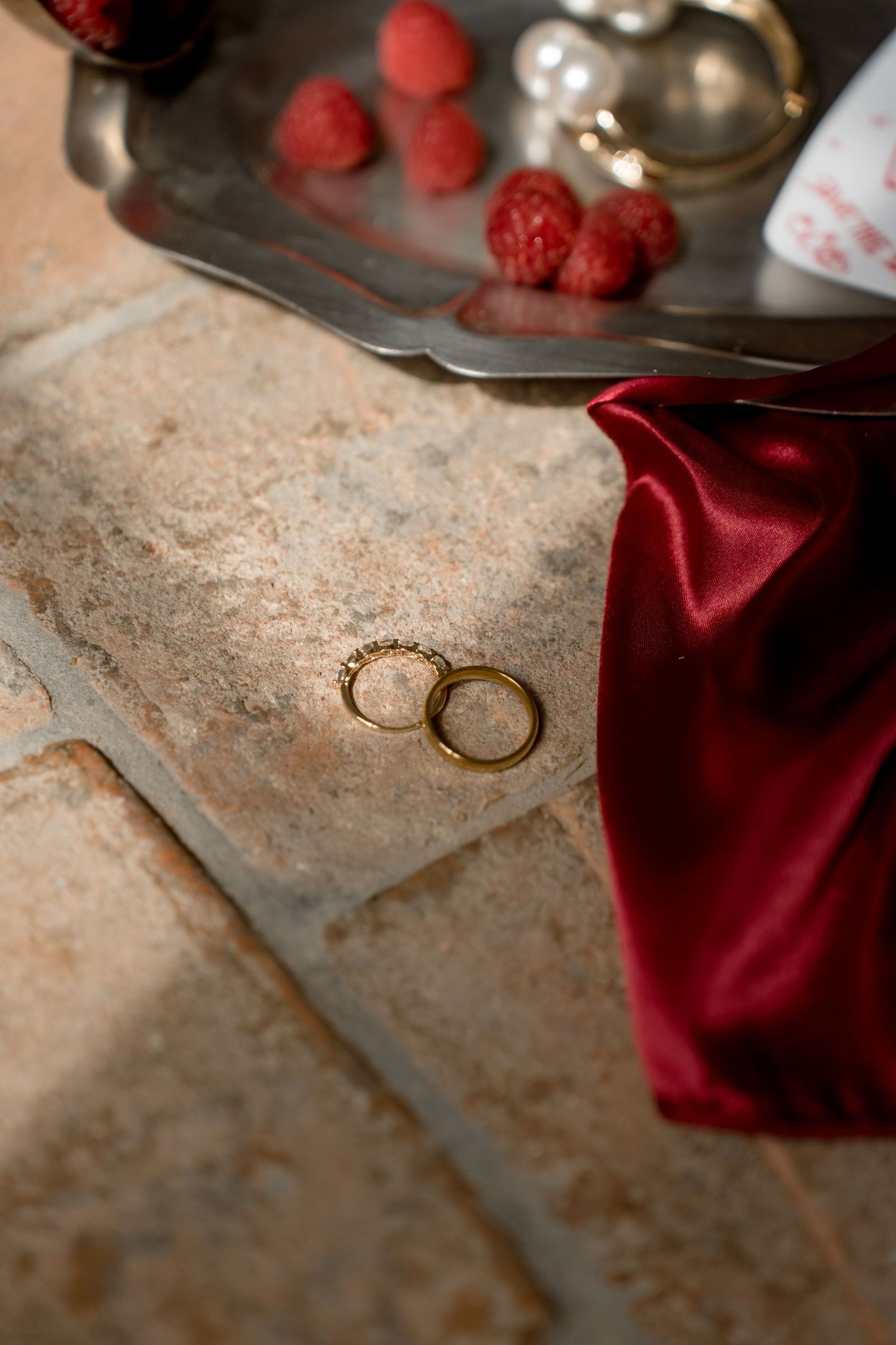 Two gold wedding rings on terracotta tiles beside red satin fabric and fresh raspberries.