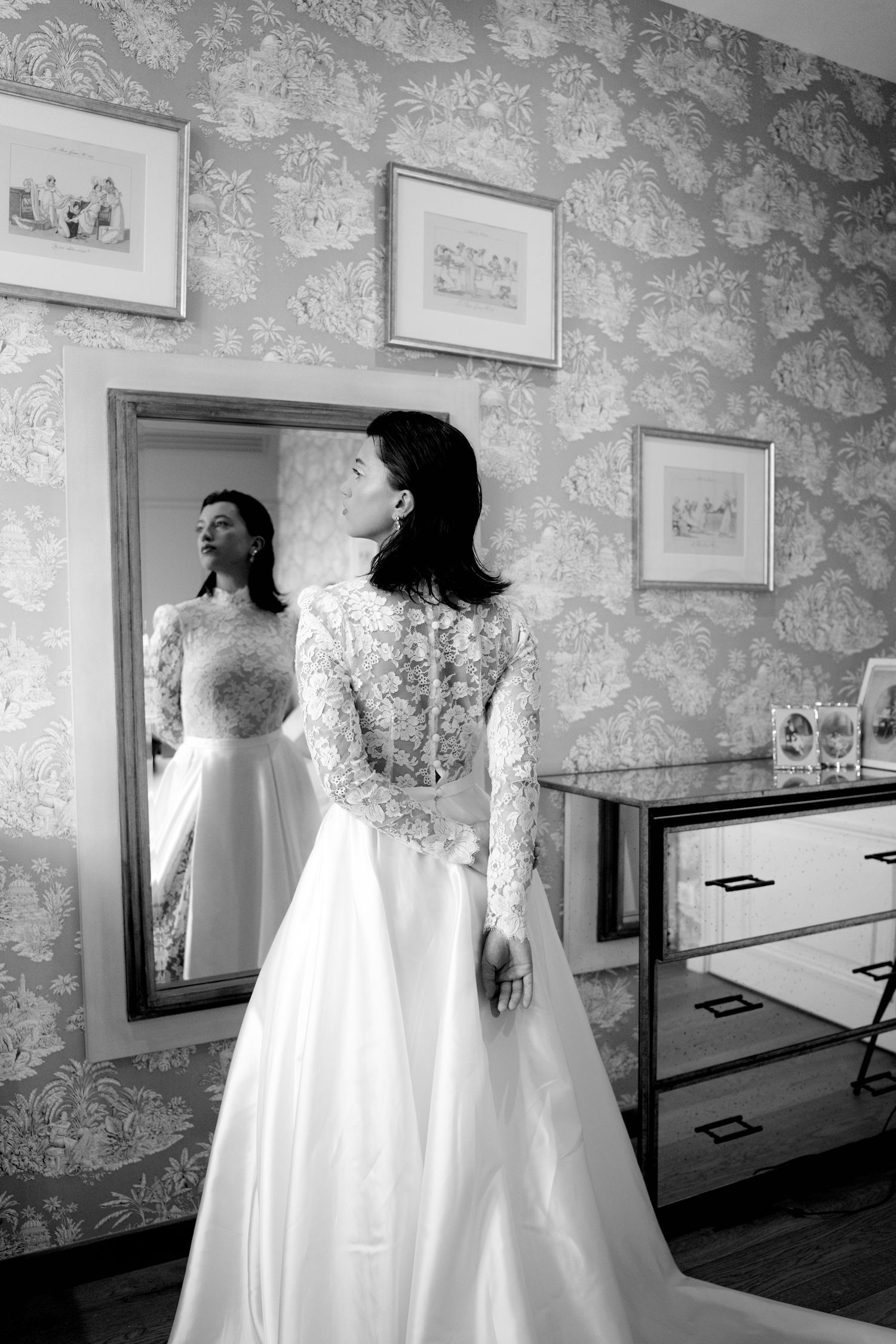 Black and white photo of bride in long-sleeve lace wedding gown admiring reflection in ornate mirror.