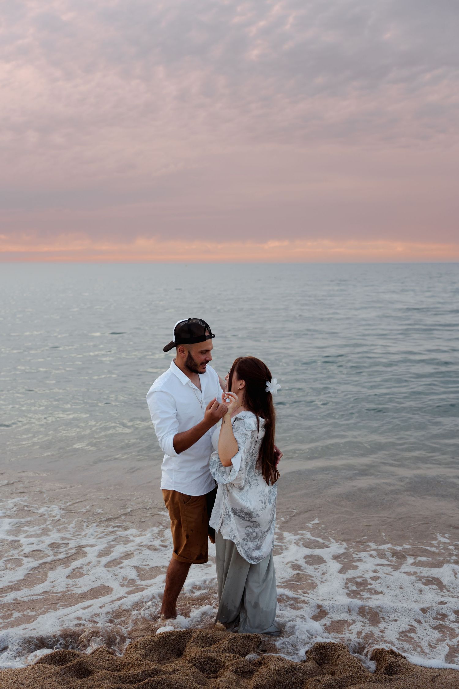 Romantic couple holding hands and facing each other on a wet beach shore at pink sunset.