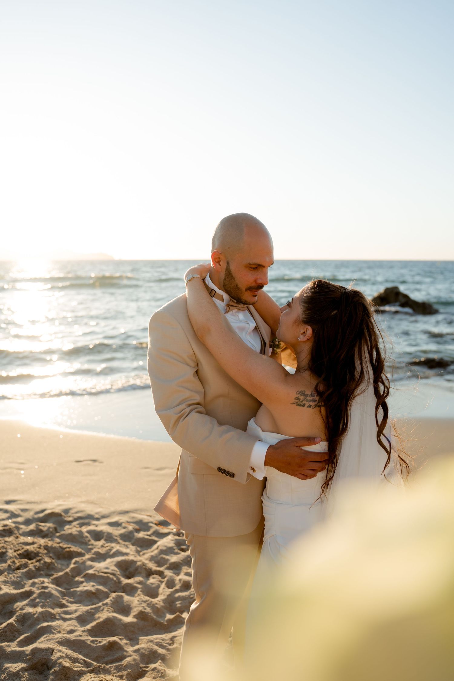 Bride and groom embracing romantically on sunlit beach with sparkling ocean waves in the background.