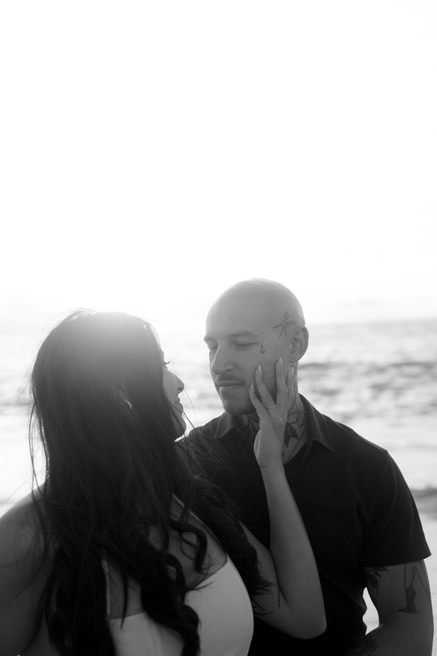 Black and white intimate portrait of a woman tenderly touching a bald mans face on a beach.
