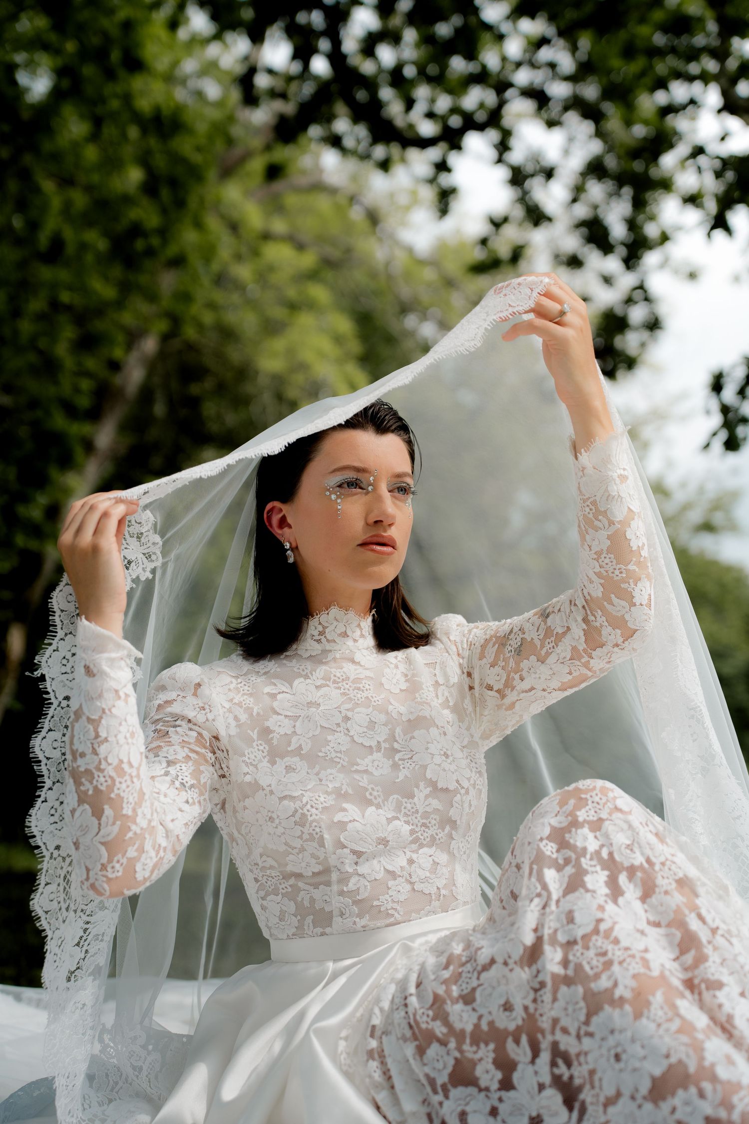 Bride in lace long-sleeve wedding gown lifting veil outdoors among green trees.