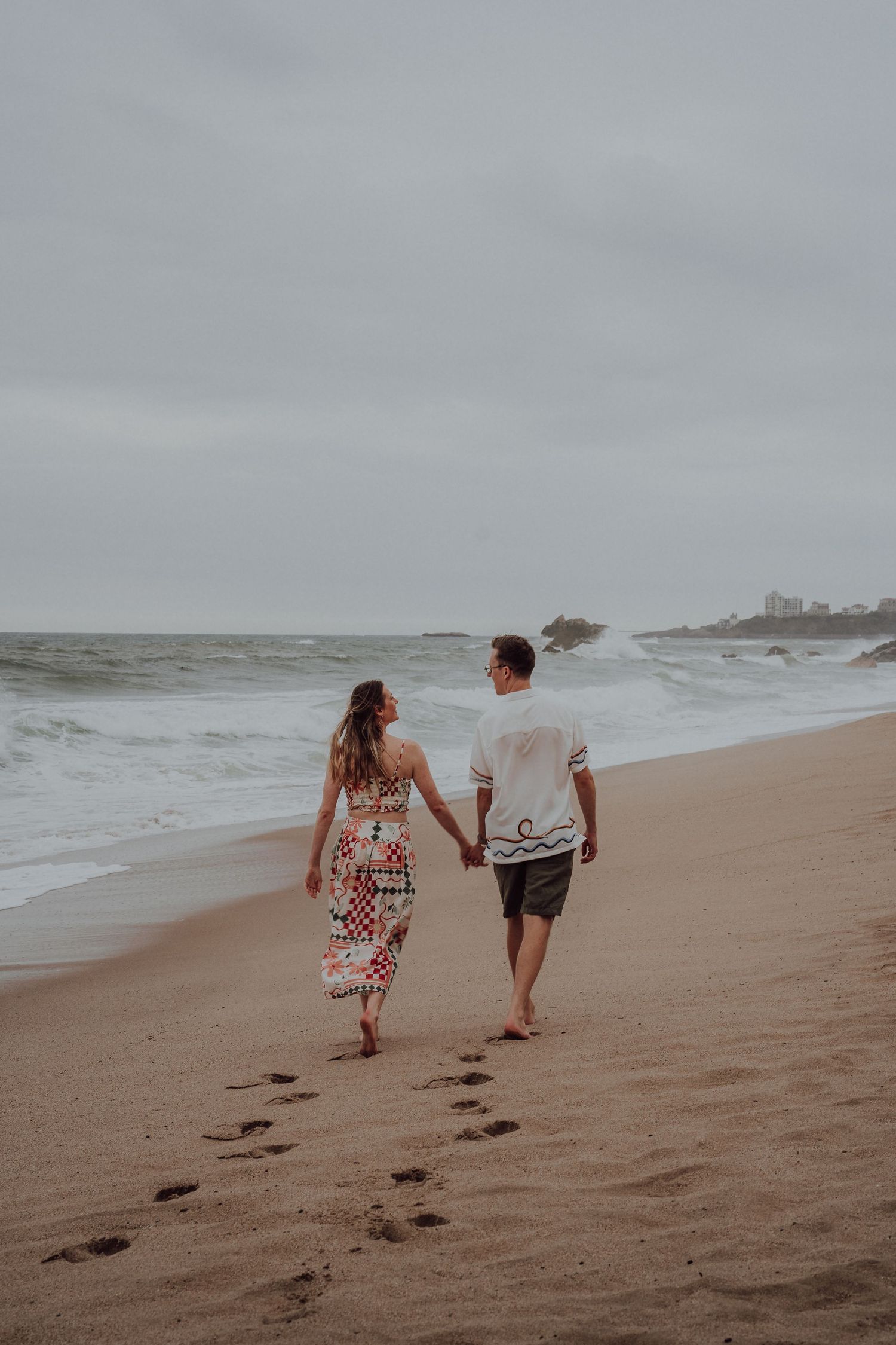 Couple holding hands walking along sandy beach with ocean waves and overcast sky.