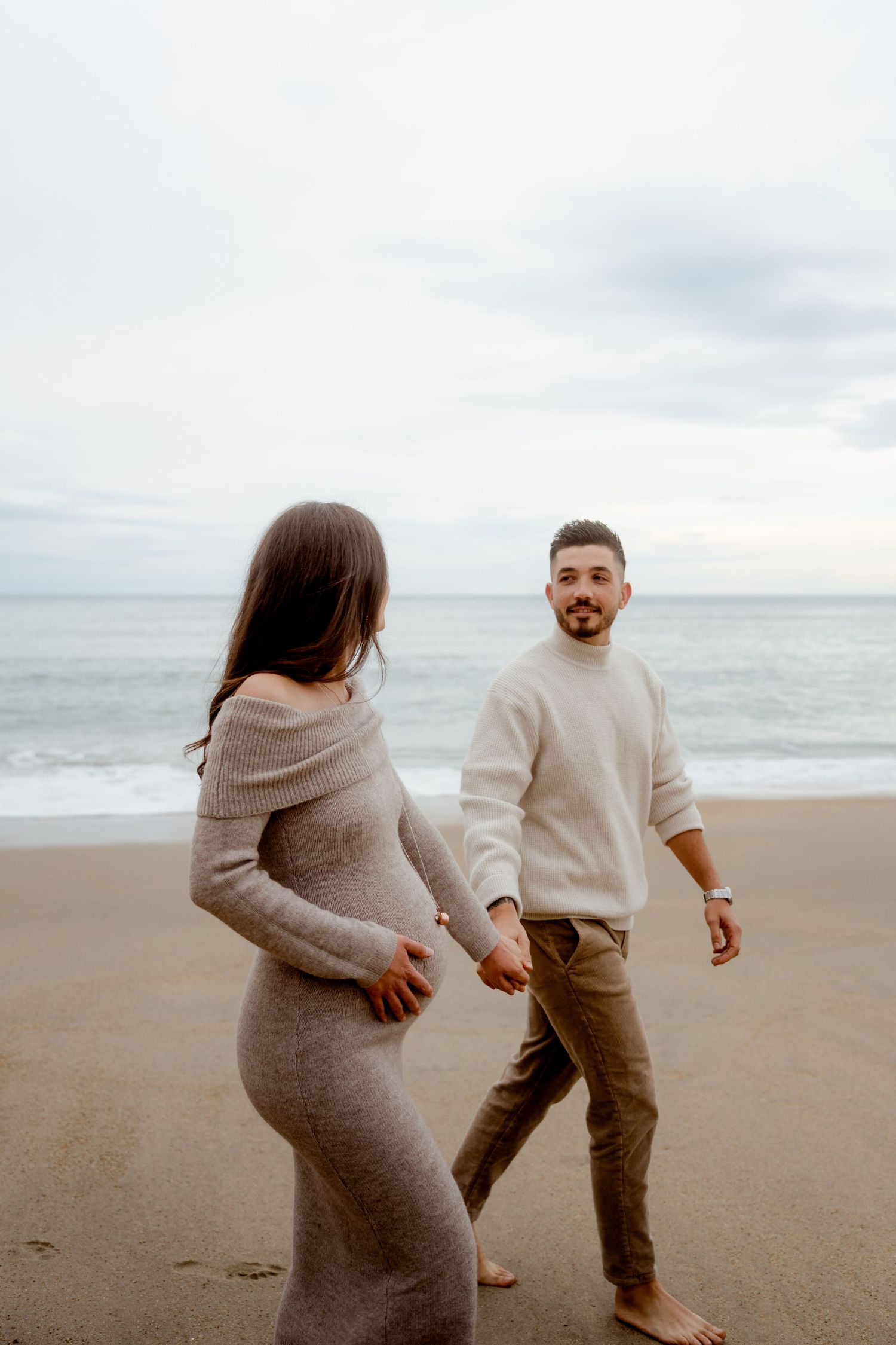 A pregnant woman in a knit dress holds hands with a man on a cloudy beach.