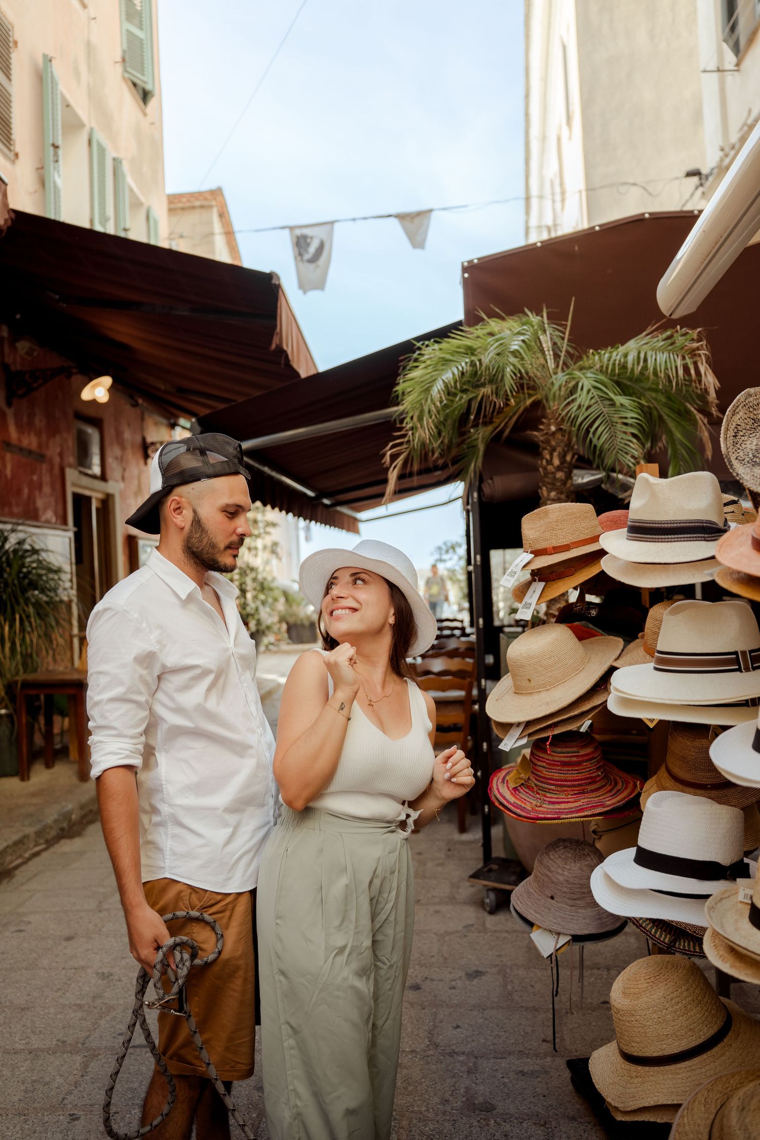 Couple smiling at each other while browsing hats at an outdoor market stall on a sunny European street.