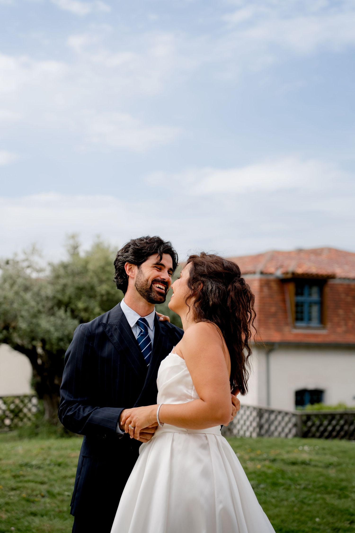 Smiling groom in navy suit gazes at bride in strapless white gown against blue sky and rustic villa backdrop.