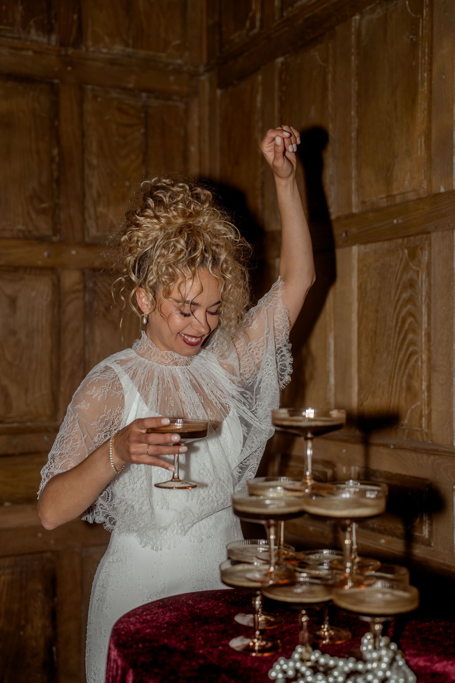 Joyful blonde bride in lace white dress dances with raised arm beside champagne tower at reception.