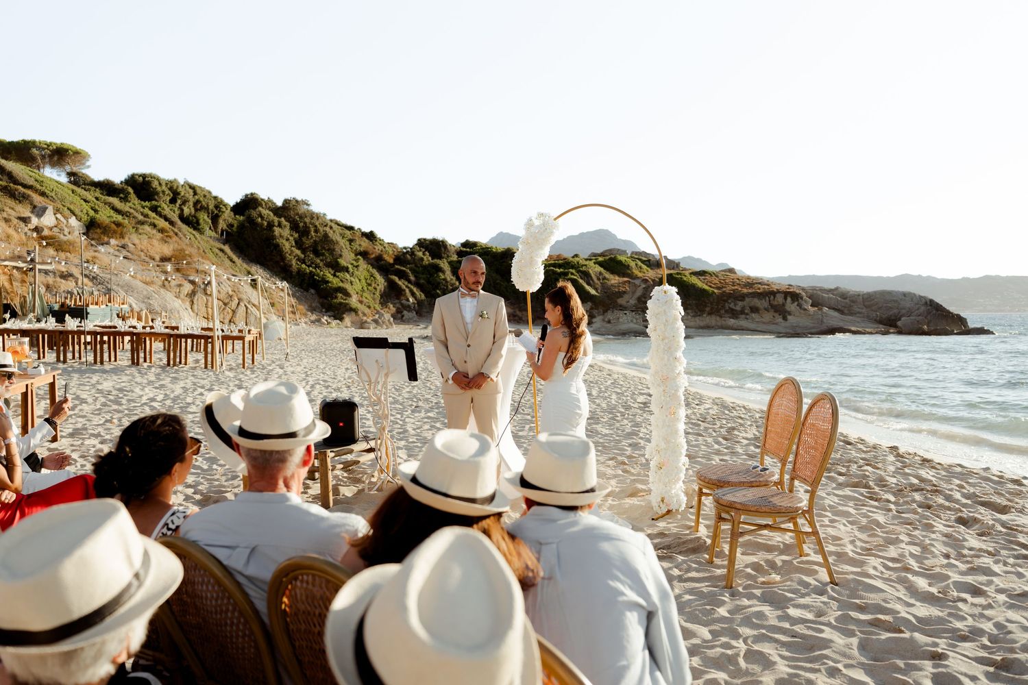 Intimate beach wedding ceremony with guests in white hats watching couple exchange vows at floral arch by the sea.