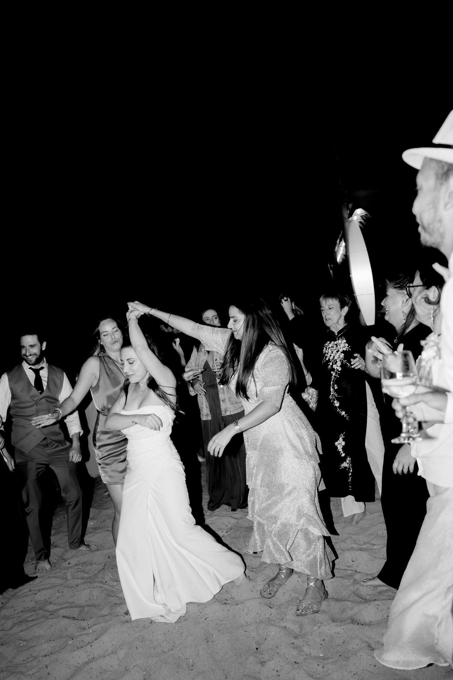 Black and white photo of bride dancing with guests on sandy beach at wedding reception.