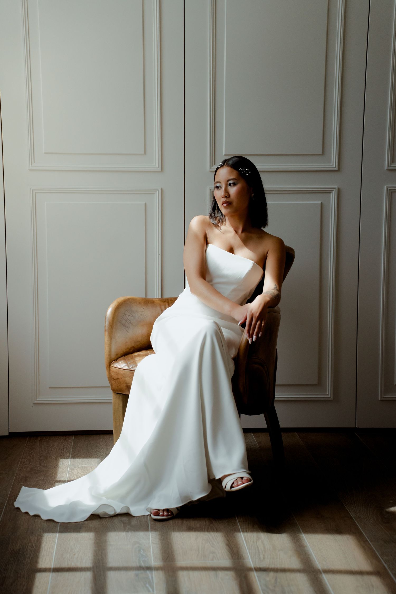 Asian bride in strapless white satin gown seated gracefully in rattan chair by paneled wall.