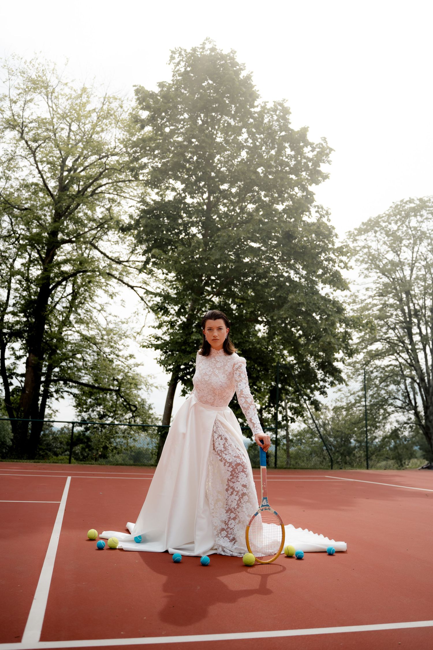 Bride in white lace wedding gown posing with vintage tennis racket on red clay court outdoors.