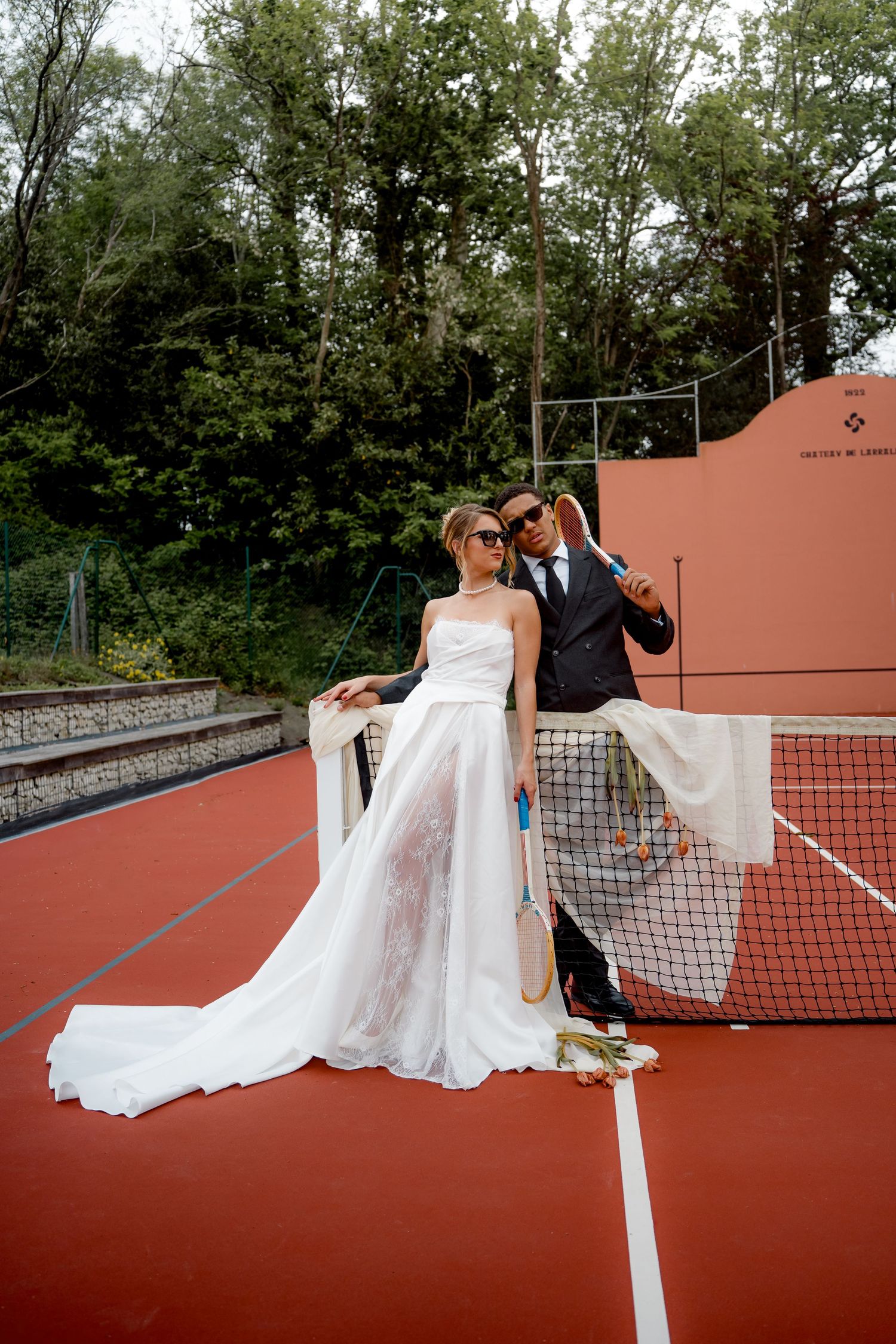 Bride and groom pose together at a tennis court net, bride wearing a flowing white gown with a long train.