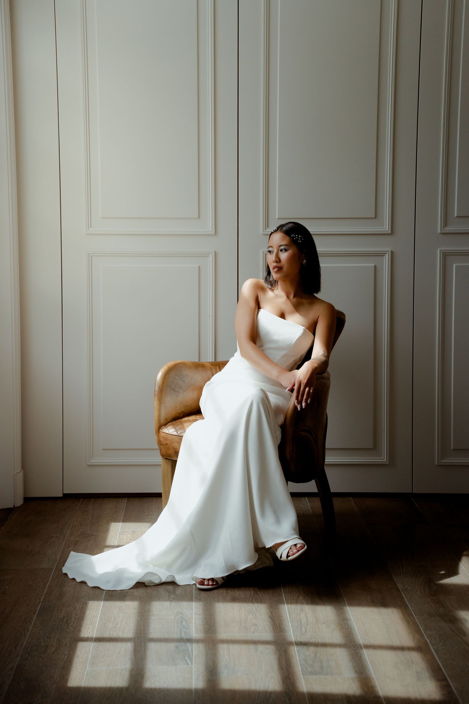 Elegant bride in minimalist white strapless gown seated in rattan chair bathed in soft natural light.