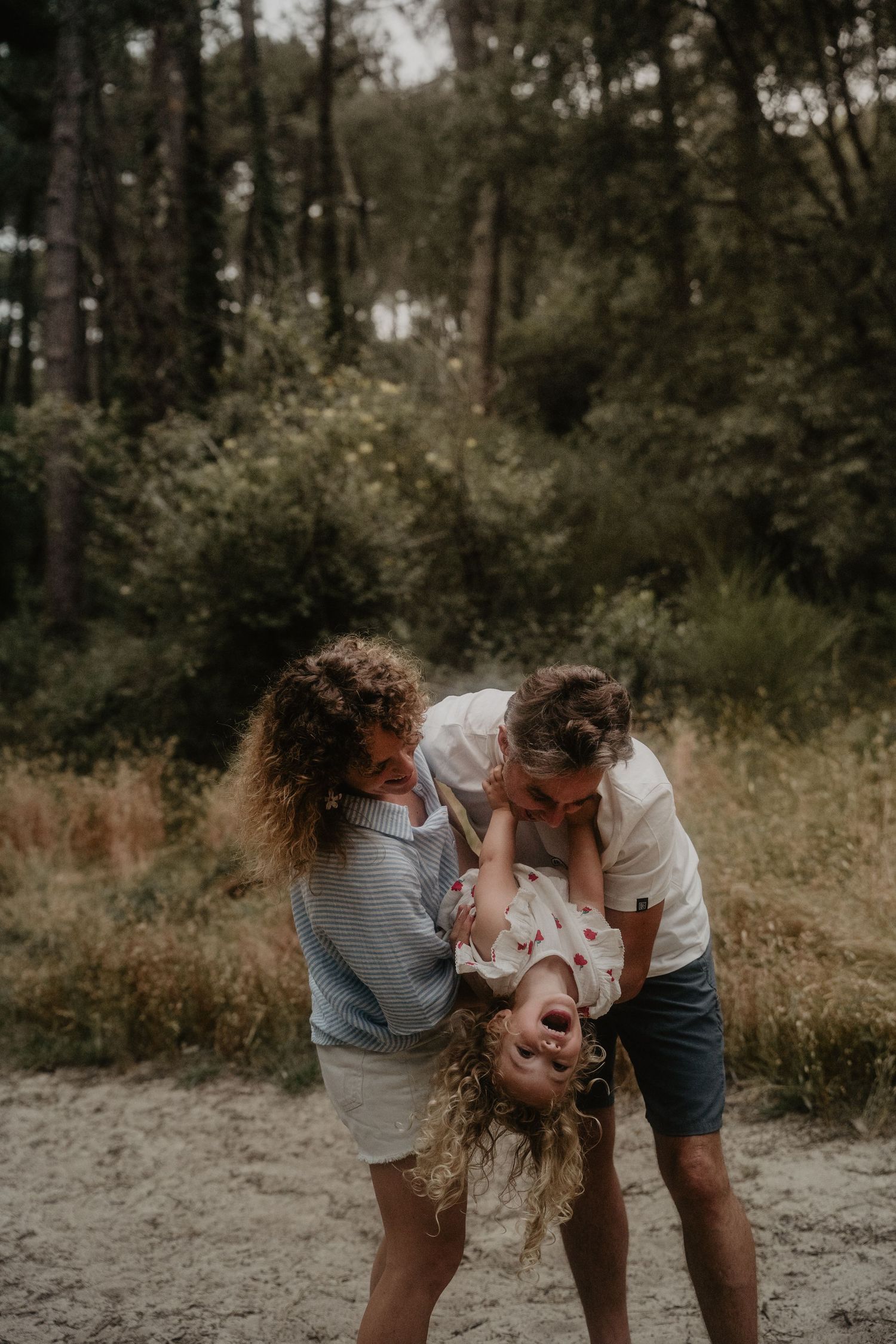 Two parents playfully hold their laughing child upside down on a sandy forest path surrounded by green trees.