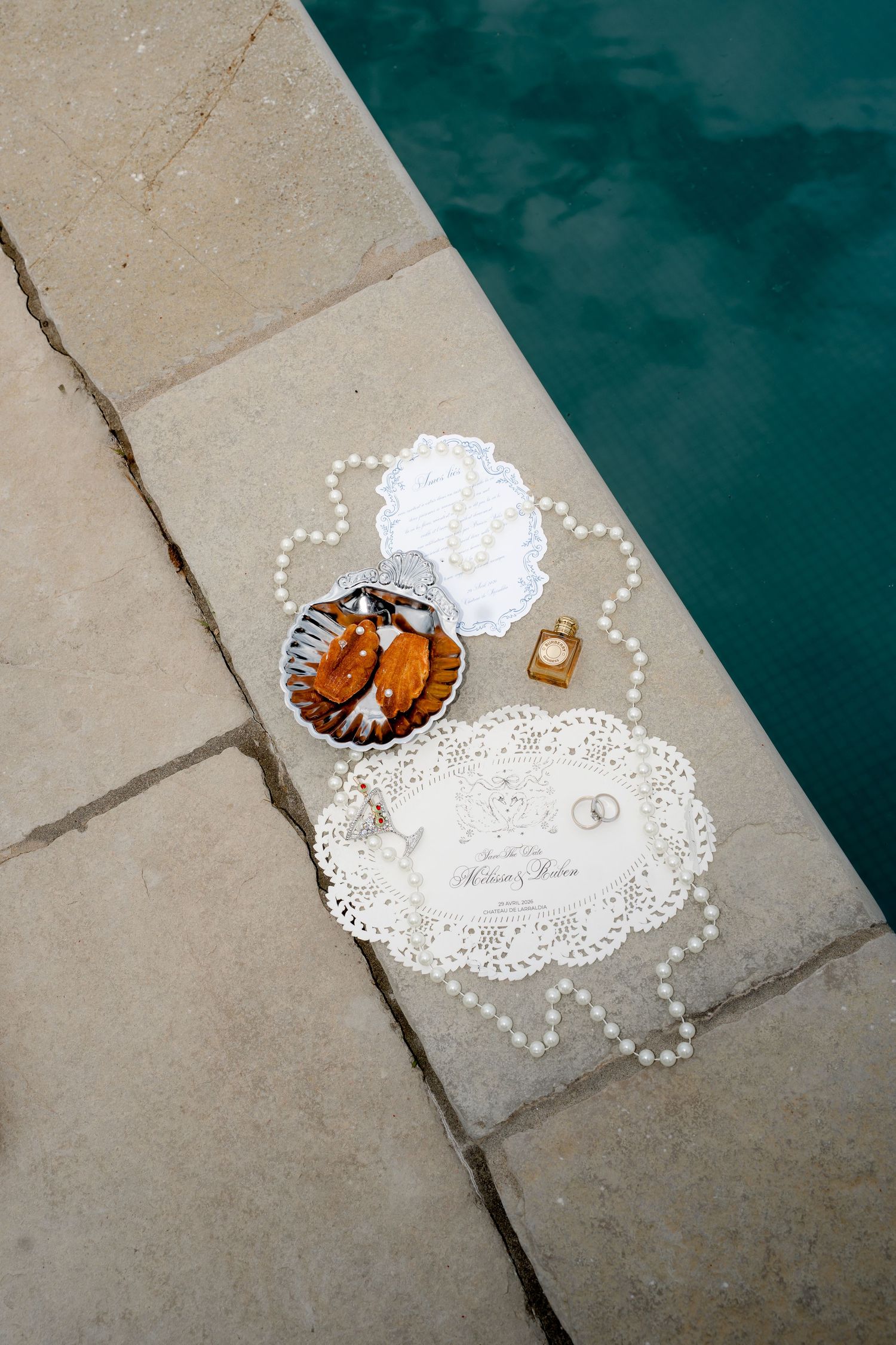 Wedding flat lay with invitation, pearl necklace, rings, and perfume on stone ledge beside turquoise pool.