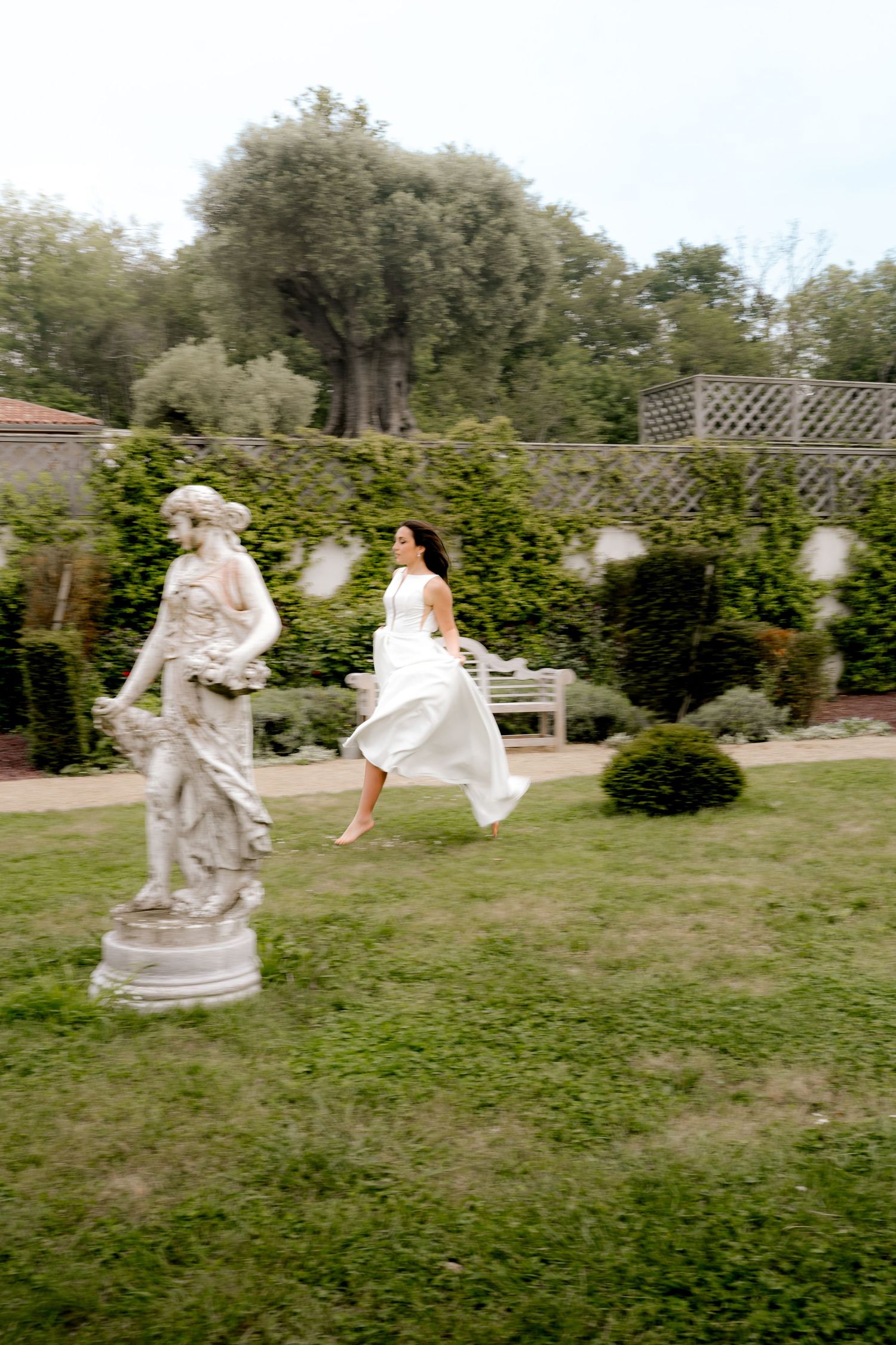 Bride in white dress running barefoot past classical stone statue in lush formal garden.