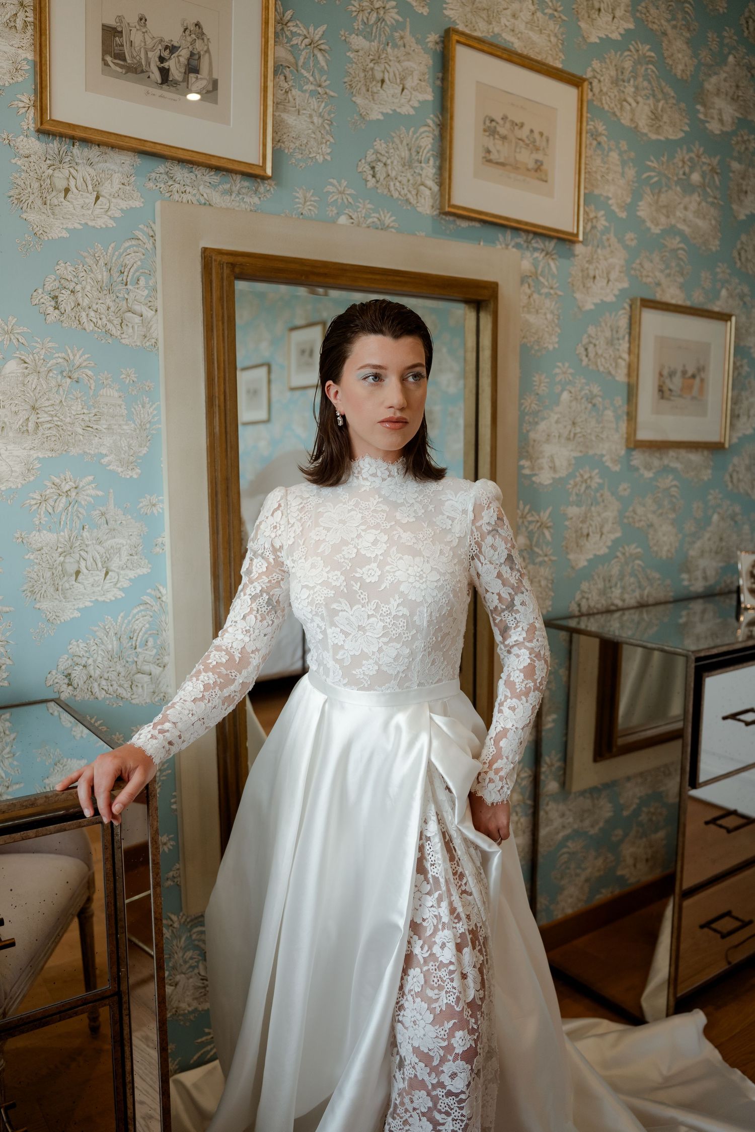 Bride in vintage-style high-neck lace wedding gown posing in room with blue floral wallpaper.