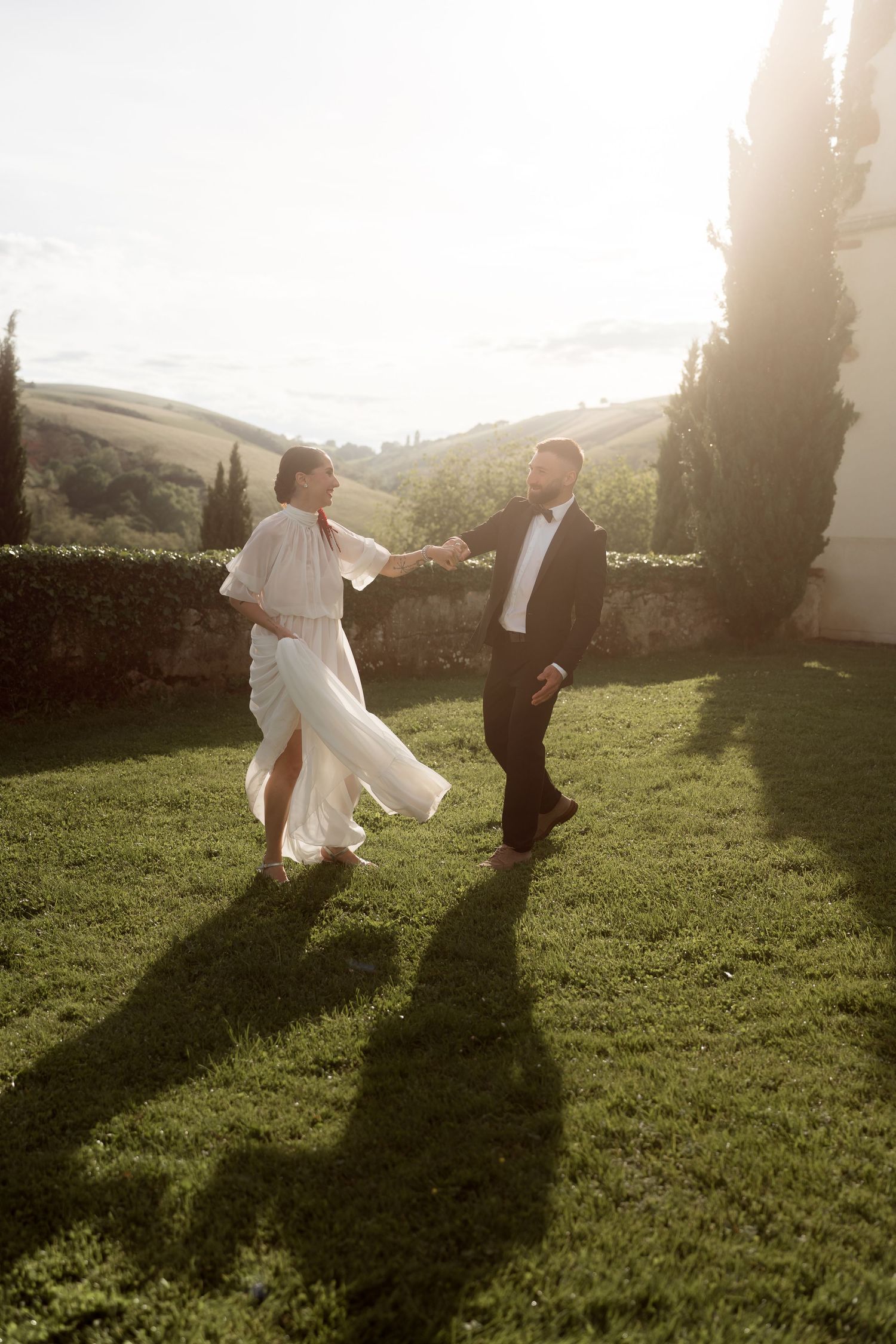 Bride and groom dancing on green lawn at golden hour with Tuscan hills backdrop.