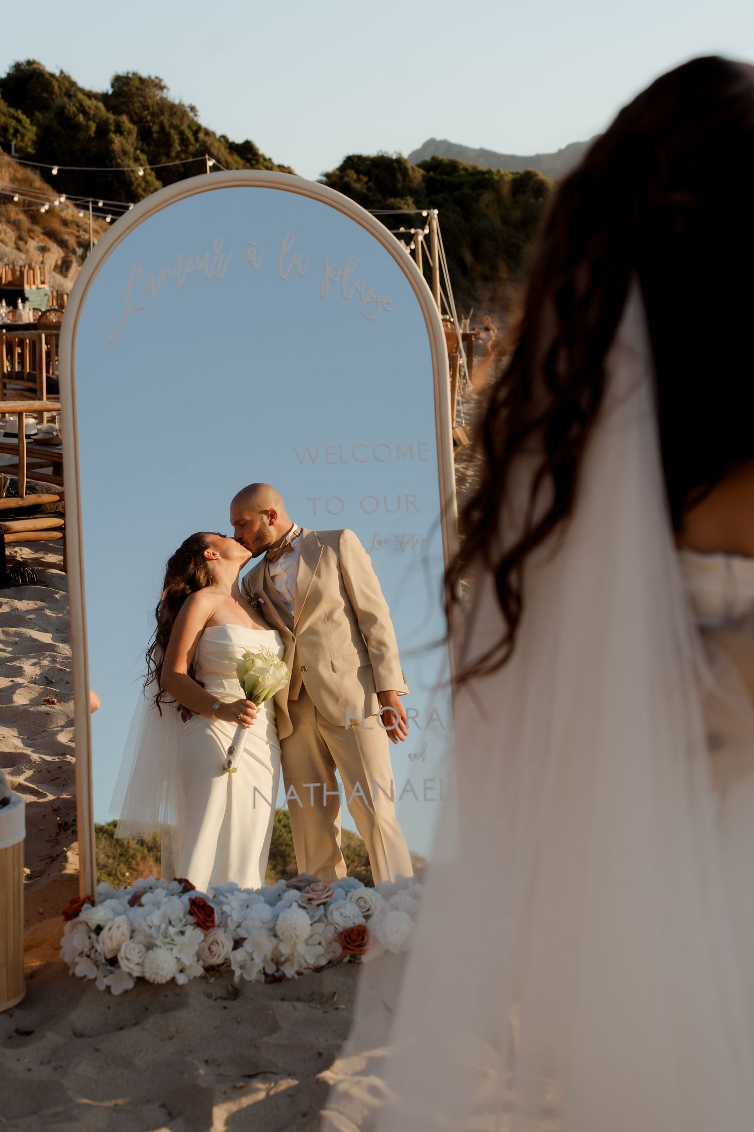 Bride and groom sharing a kiss reflected in an arched welcome mirror at a beach wedding ceremony.