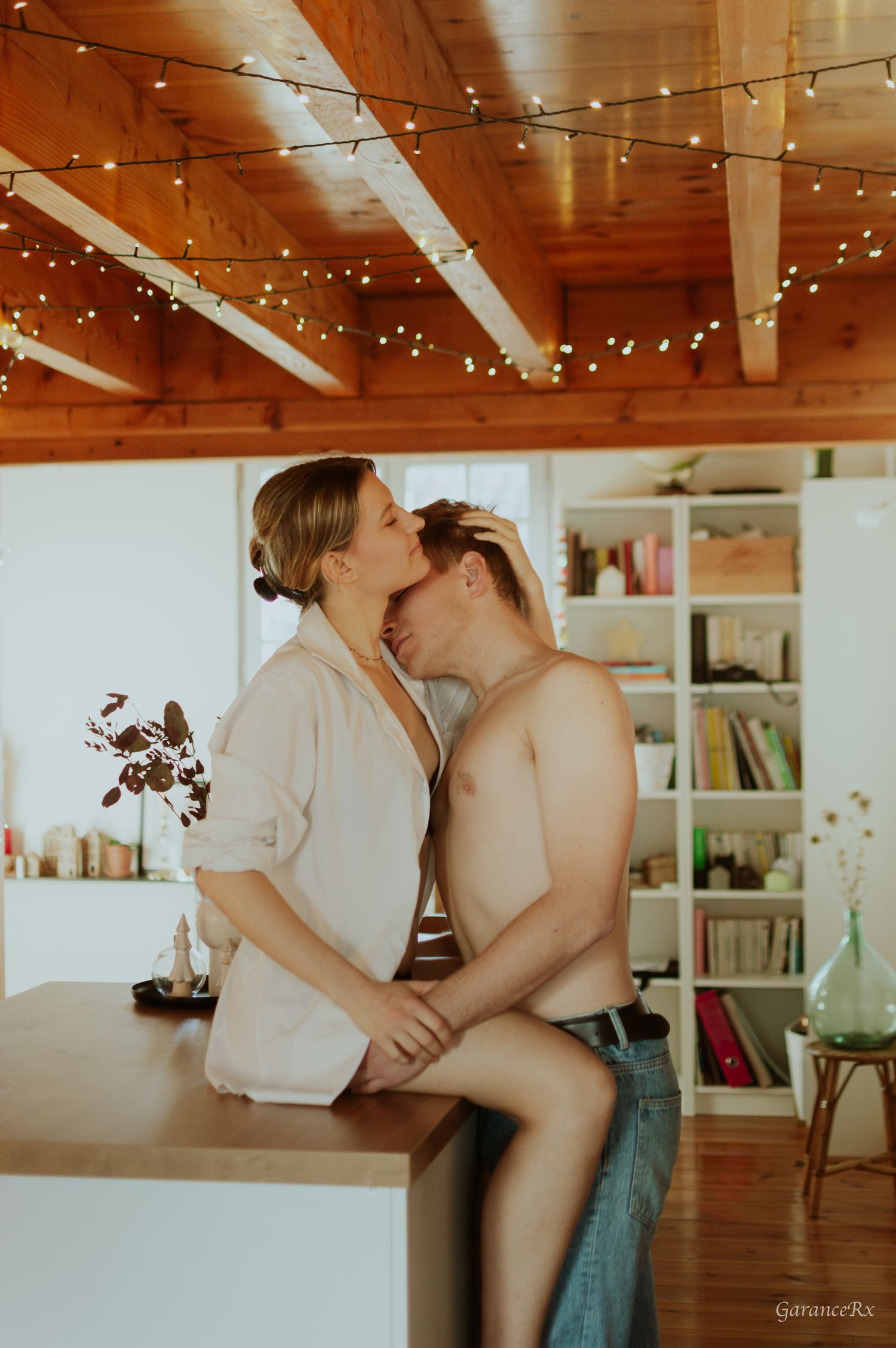 Couple sharing a romantic kiss on a kitchen counter with warm fairy lights and wooden beamed ceiling.