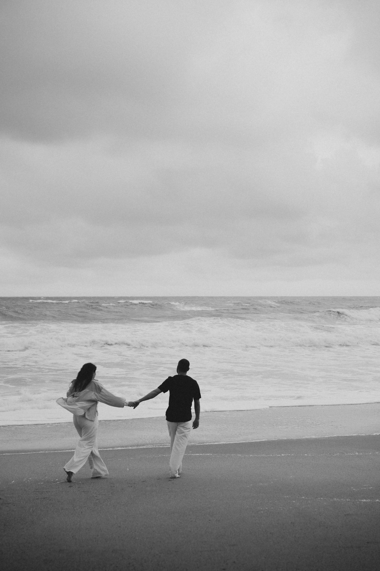 Black and white image of a couple holding hands walking toward crashing ocean waves on a beach.