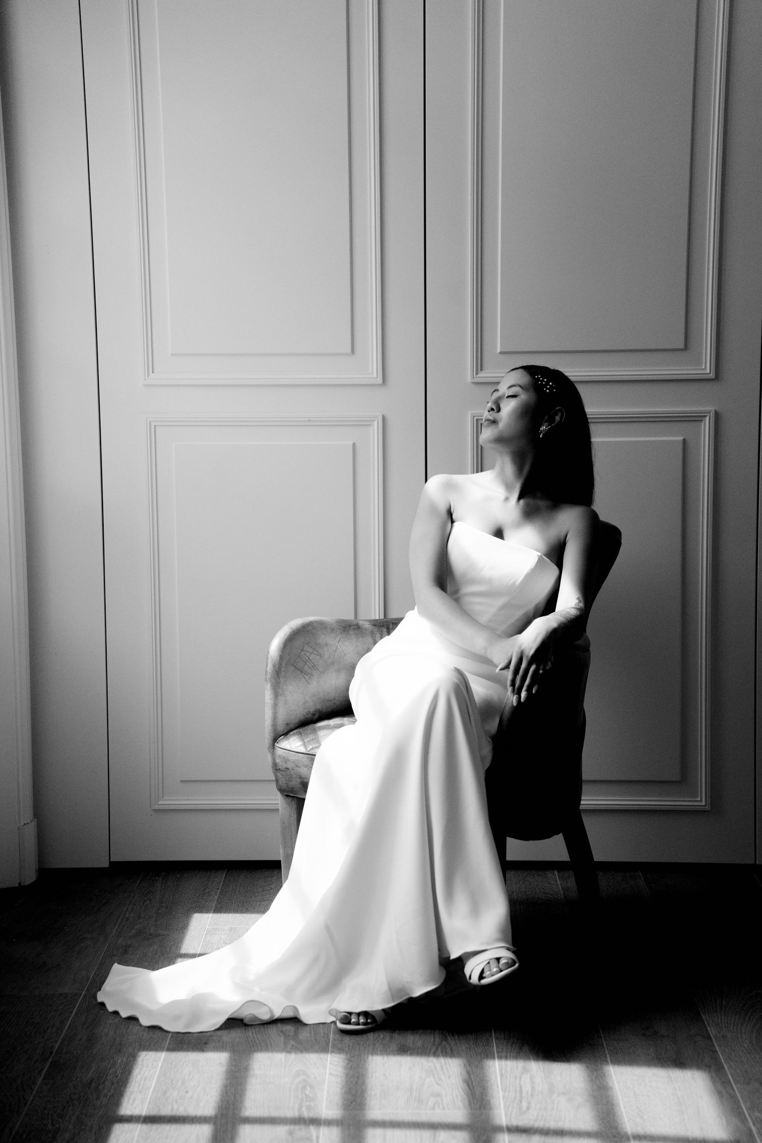 Black and white portrait of bride in strapless satin gown seated in chair with dramatic window light.