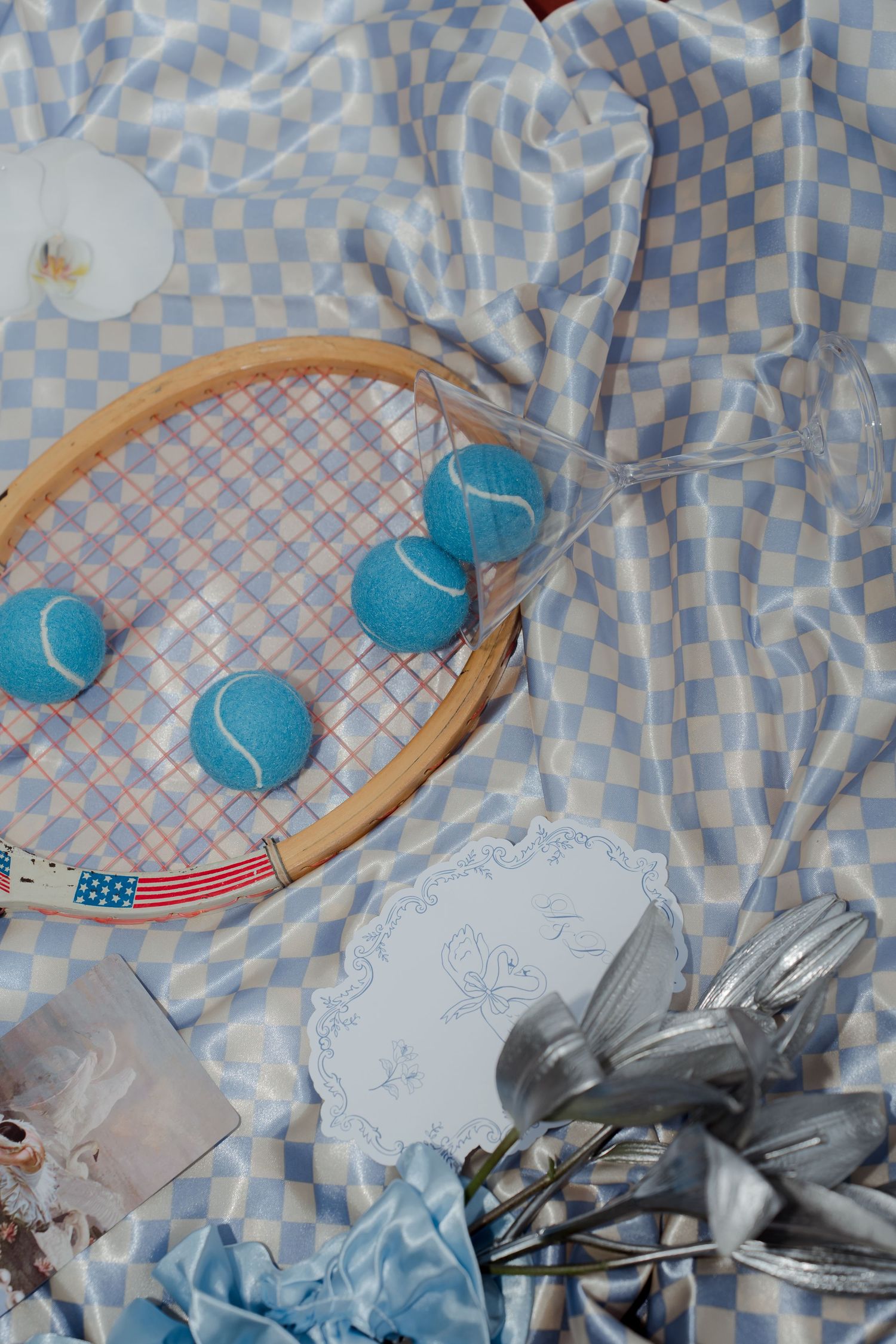 Close-up of vintage tennis racket with blue balls, lace doily, and silver ribbons on checkered fabric.