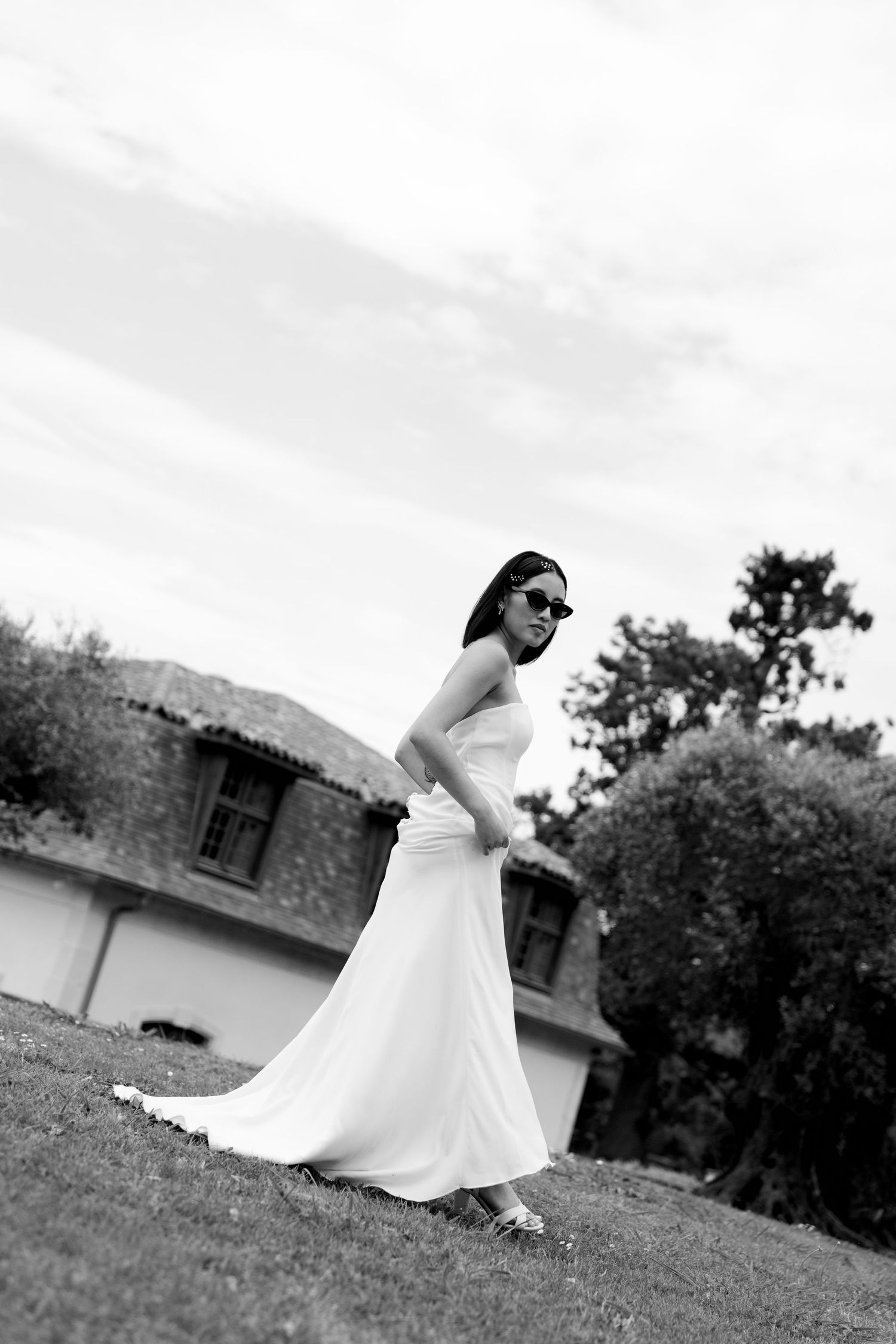 Black and white photo of bride in flowing white dress and sunglasses posing on lawn near rustic building.