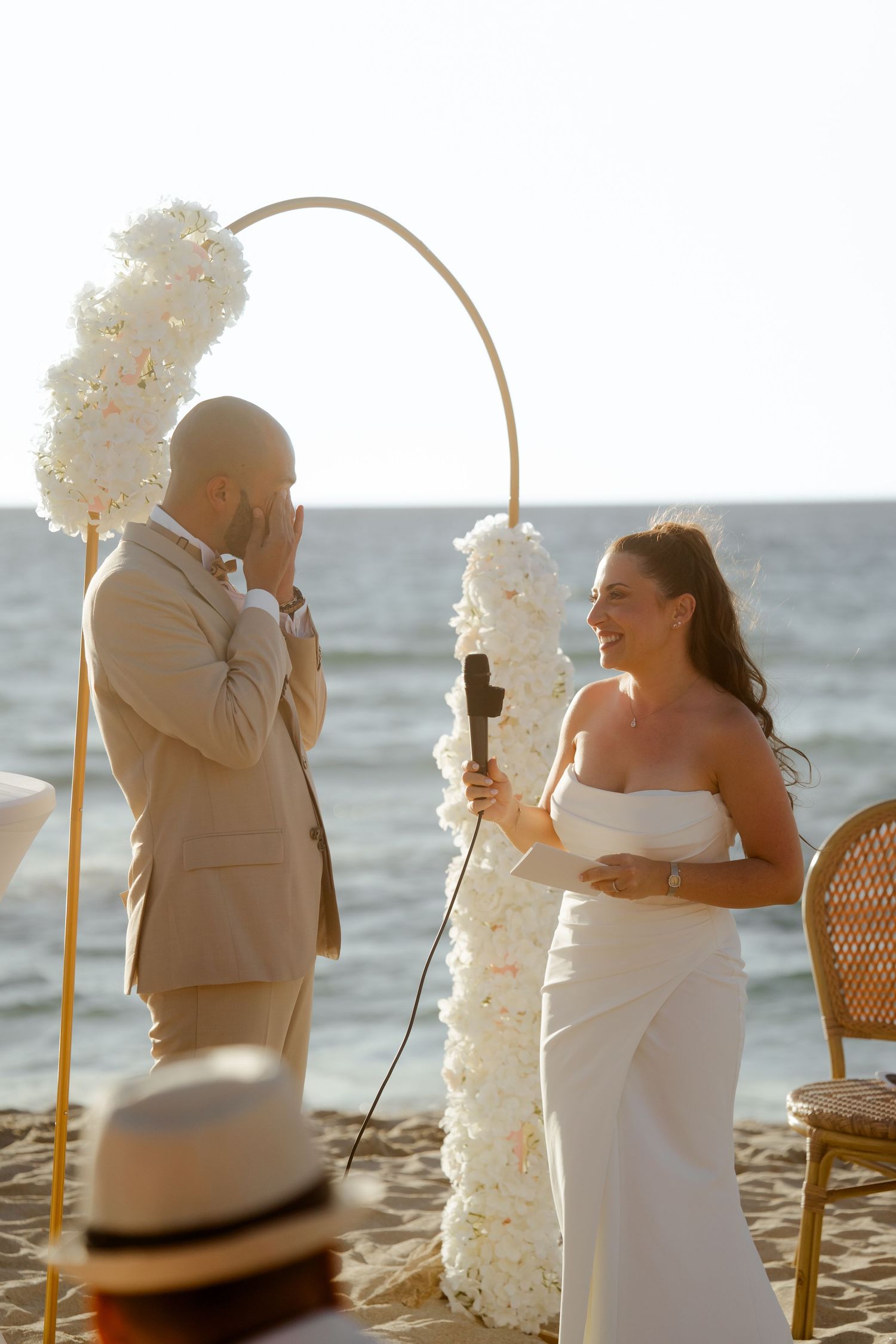 Emotional groom covers face as smiling bride reads heartfelt vows at sunset beach wedding ceremony.