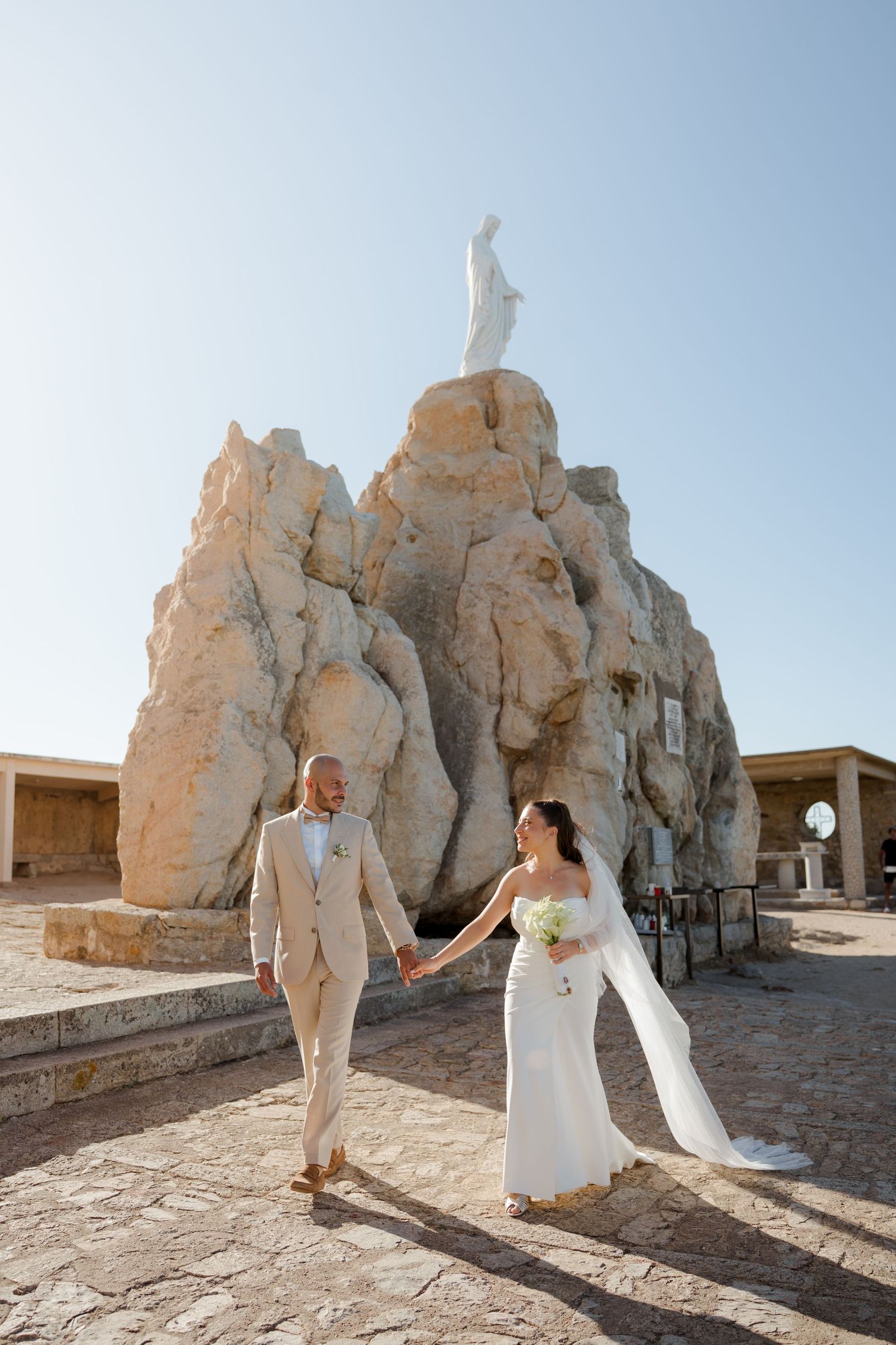 Bride and groom in elegant attire walk hand in hand before dramatic rocky outcrop with white religious statue.