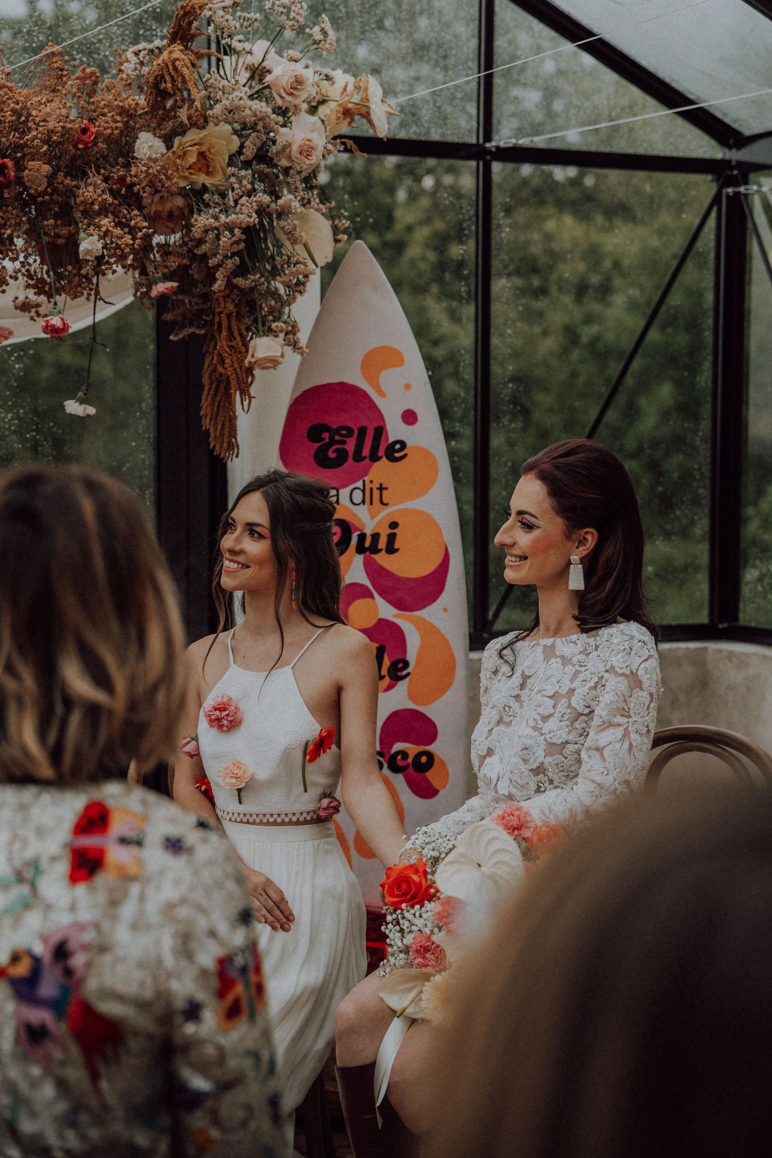 Two brides sitting together at their wedding ceremony in a greenhouse with a colorful retro disco sign.