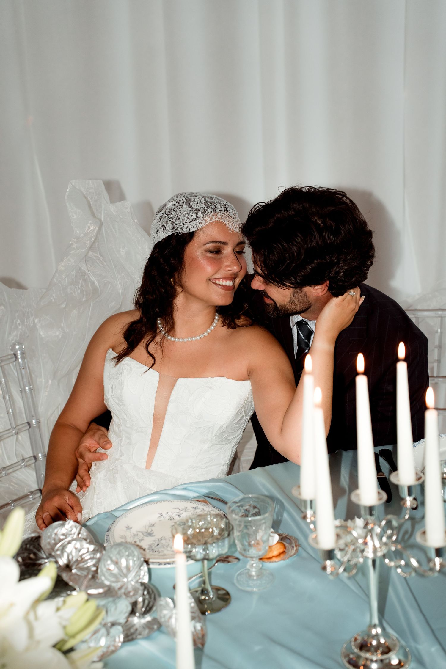 Smiling bride in pearl necklace and groom share intimate moment at candlelit wedding reception table.
