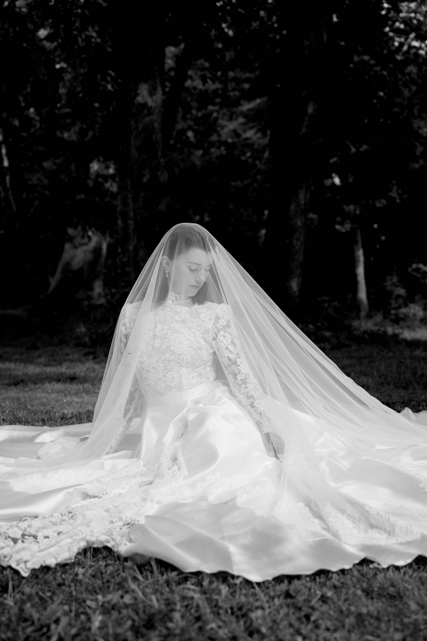 Black and white photo of a bride sitting outdoors in a flowing gown with a long veil draped over her.