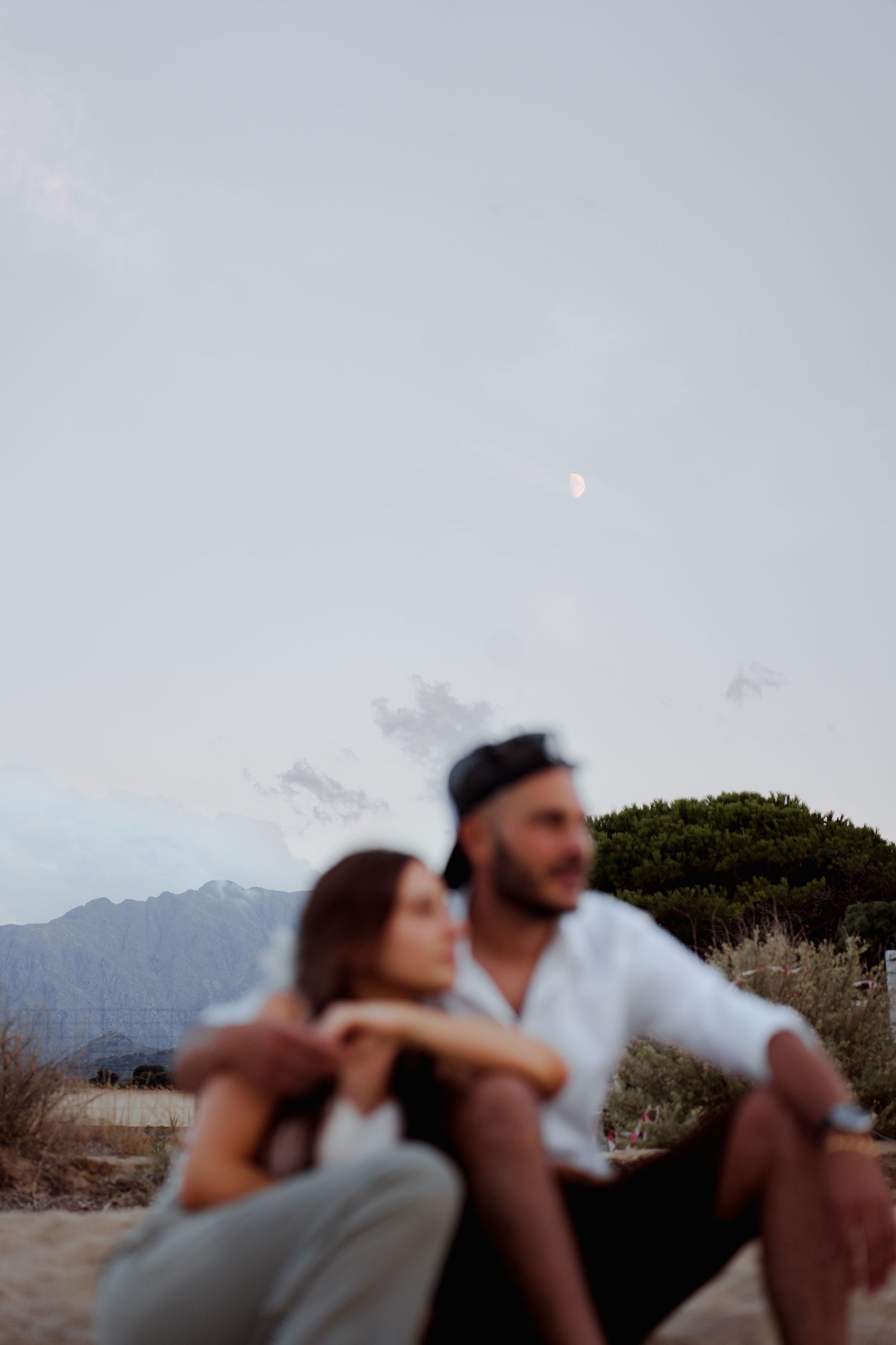 Couple sitting together outdoors at dusk with mountains and a crescent moon in the sky.