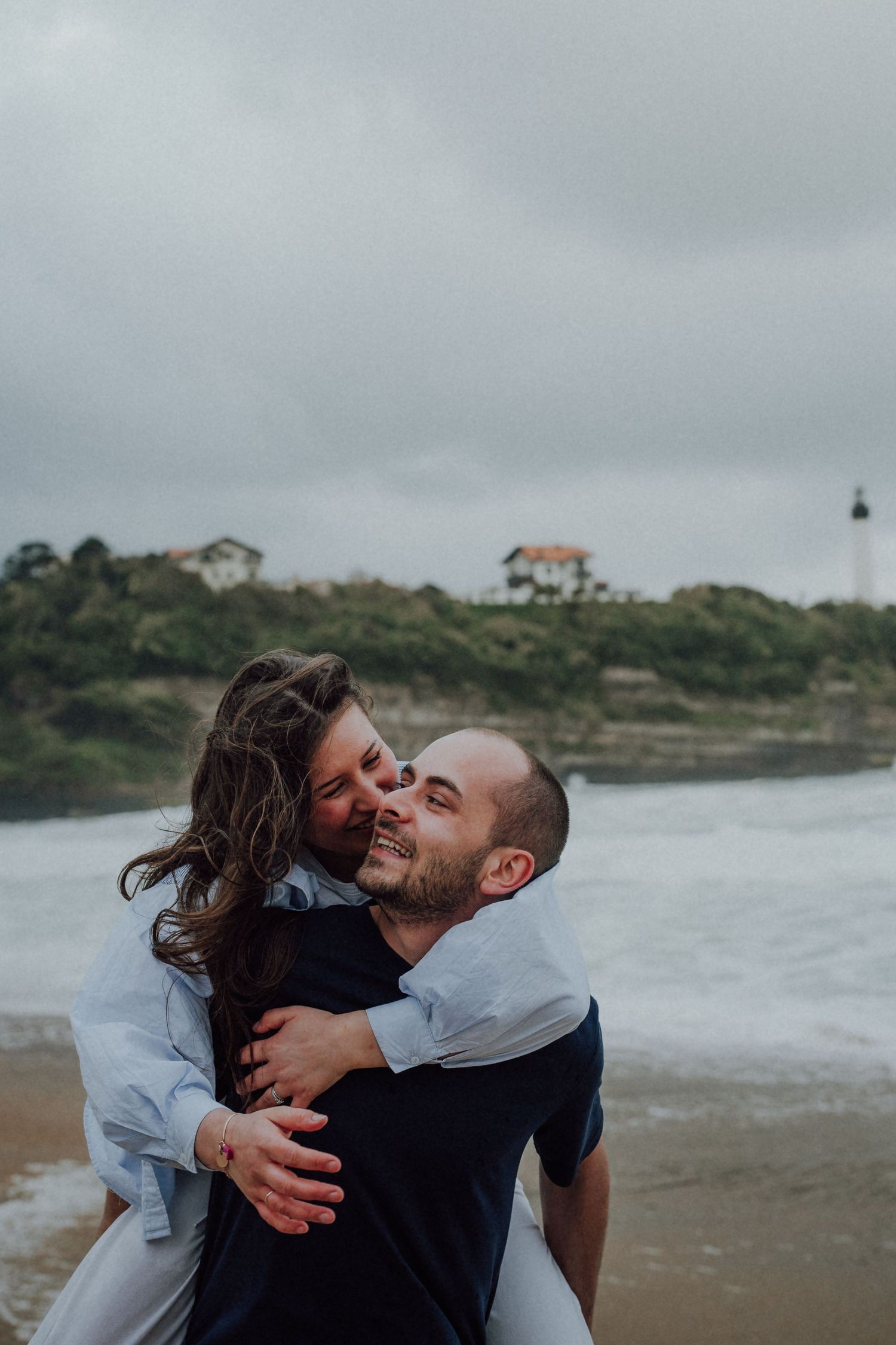 Happy couple sharing a tender piggyback hug on a beach with rocky cliffs and lighthouse in background.