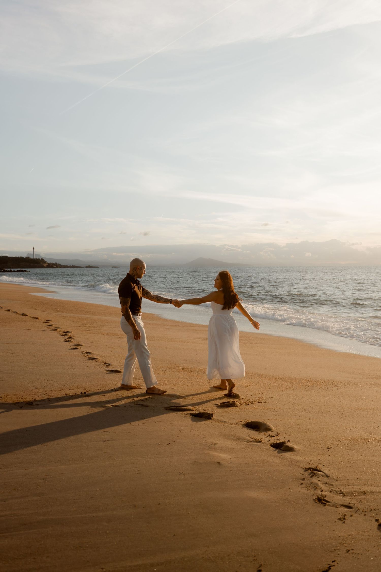 Couple holding hands and dancing on a sandy beach at sunset with footprints trailing behind them.