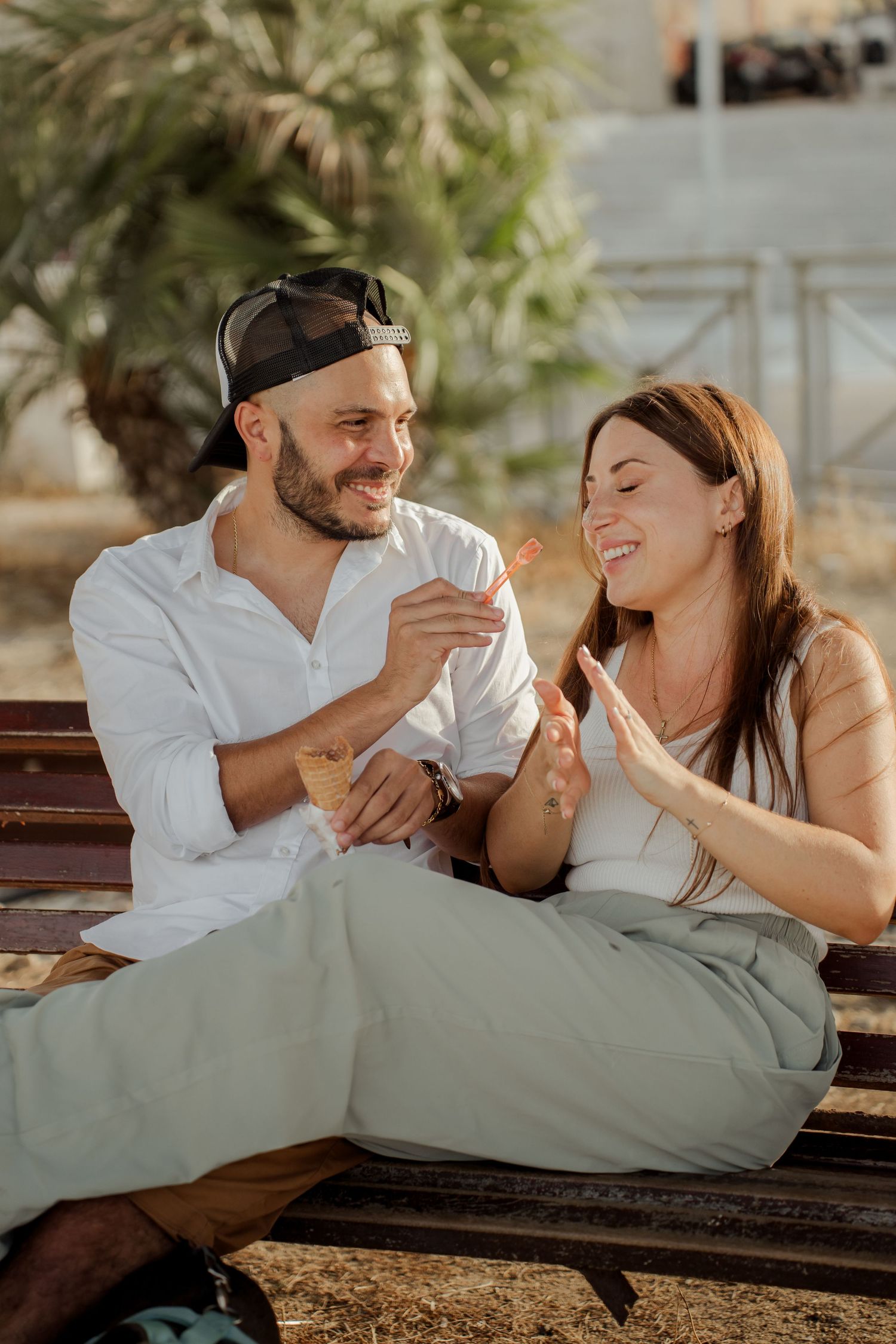 Laughing couple sharing ice cream cones while sitting together on a bench in warm sunlight.