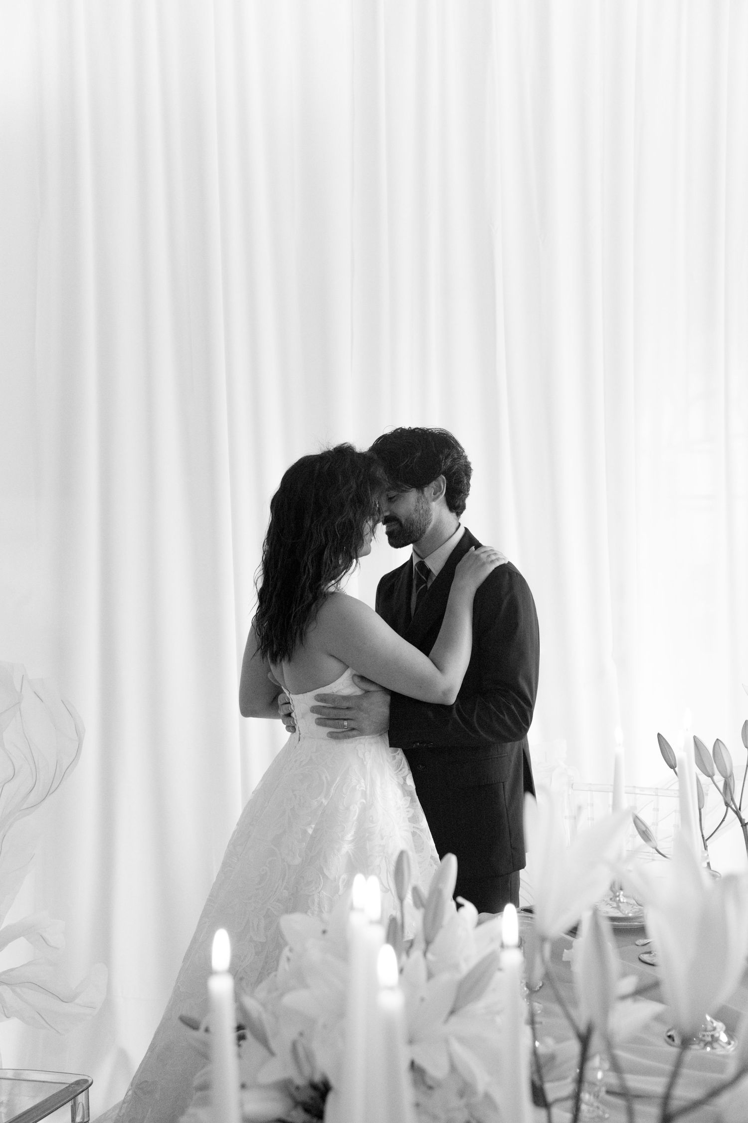 Bride and groom sharing a romantic embrace near candles and white lilies in black and white.