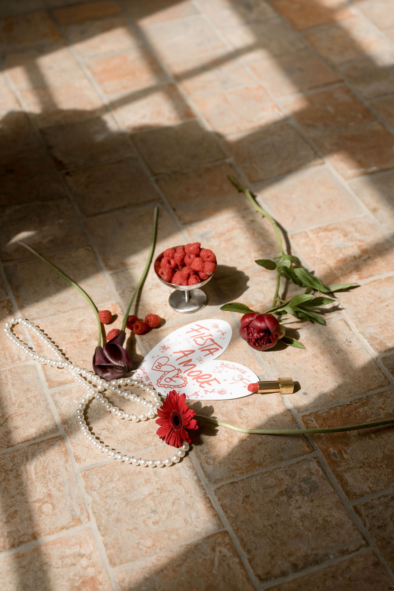 Flat lay of red roses, raspberries, pearl necklace, lipstick, and calligraphy card on sunlit tile floor.
