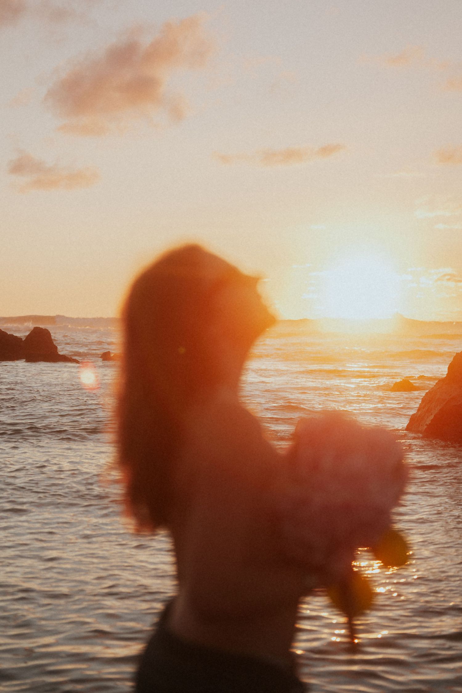 Blurred silhouette of a woman holding flowers against a vivid ocean sunset.