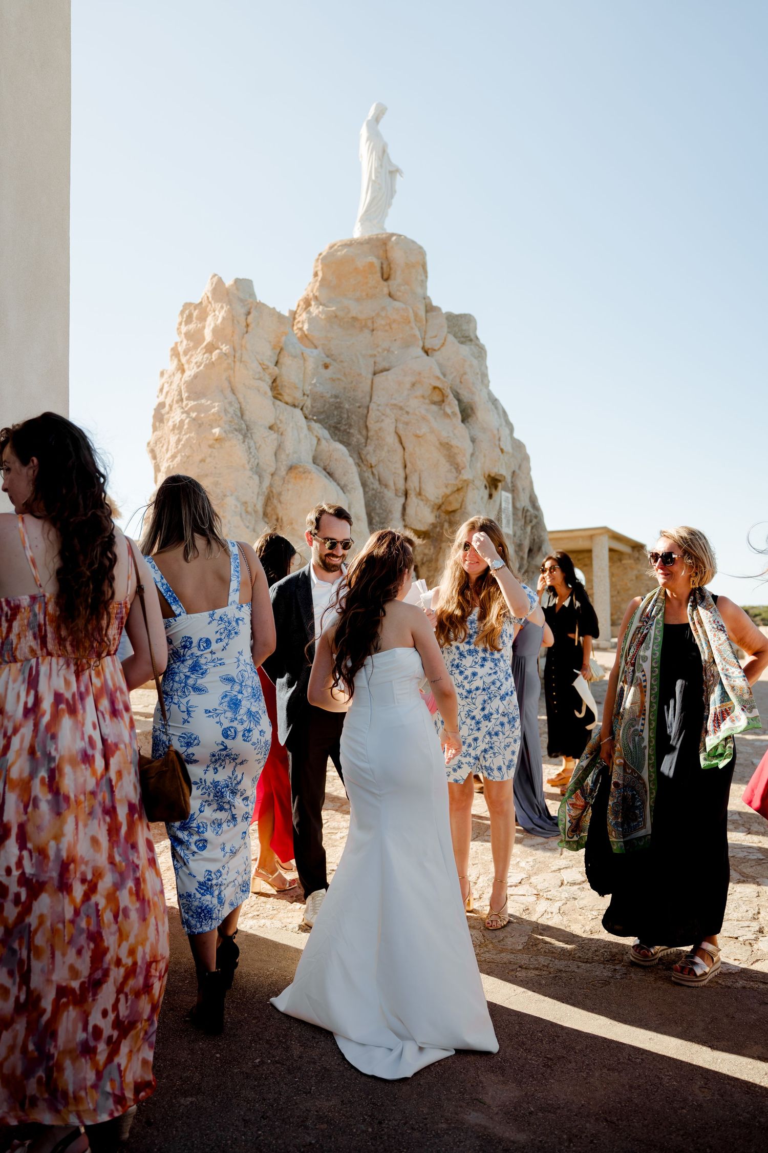 Bride in white dress mingling with wedding guests near rocky statue monument in sunny setting.