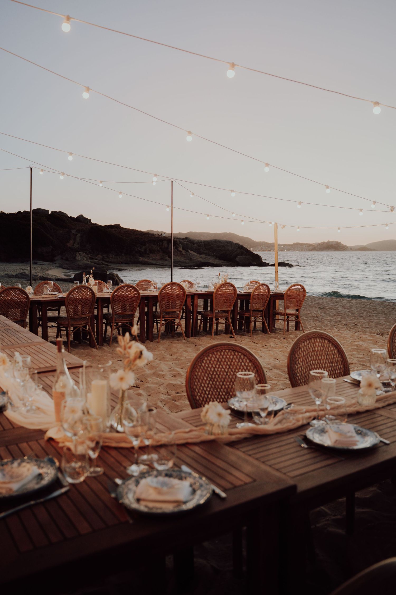 Romantic beach wedding reception at dusk with string lights, rattan chairs, and ocean backdrop.