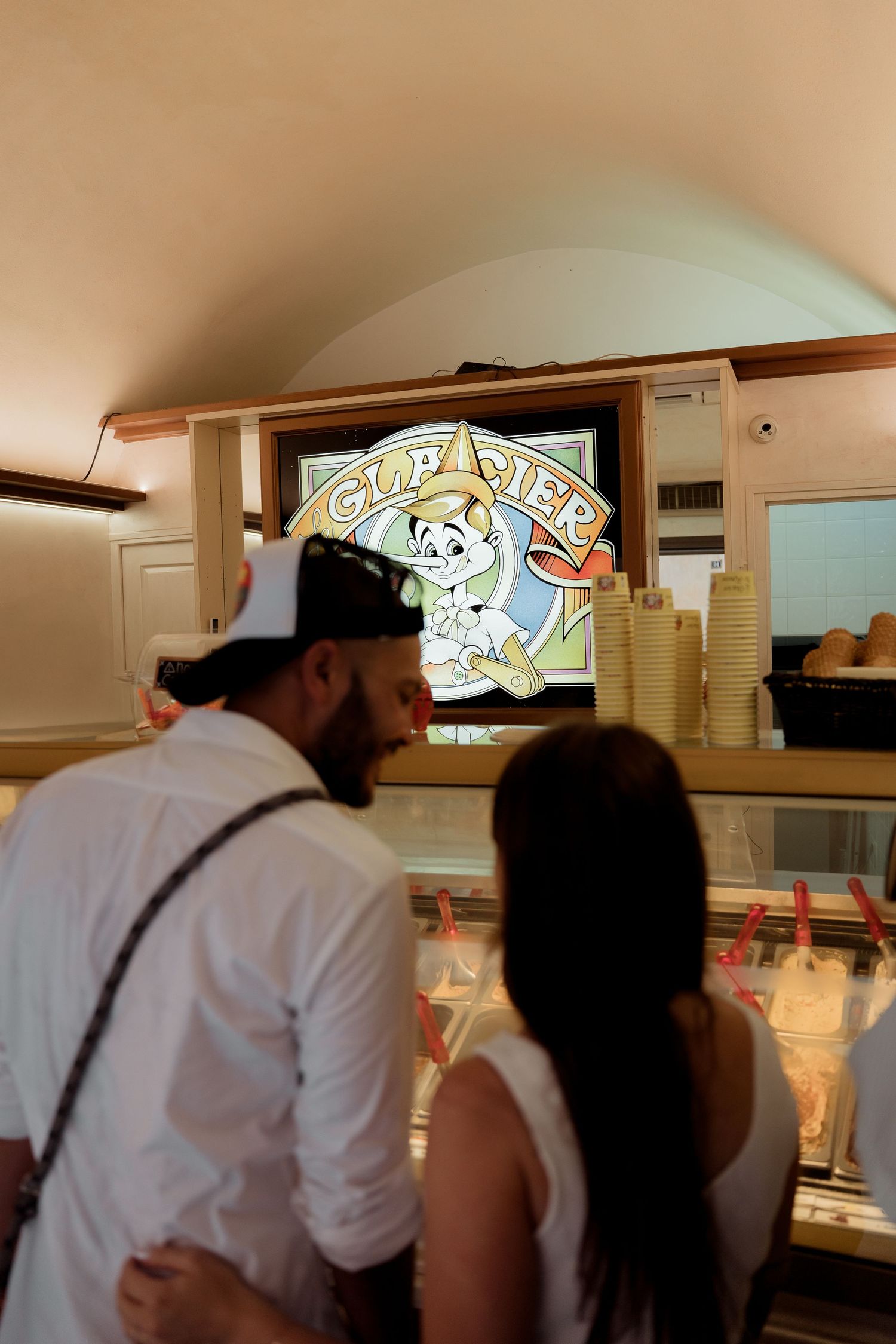 Couple ordering ice cream at a gelato shop counter with a colorful cartoon sign above the display.