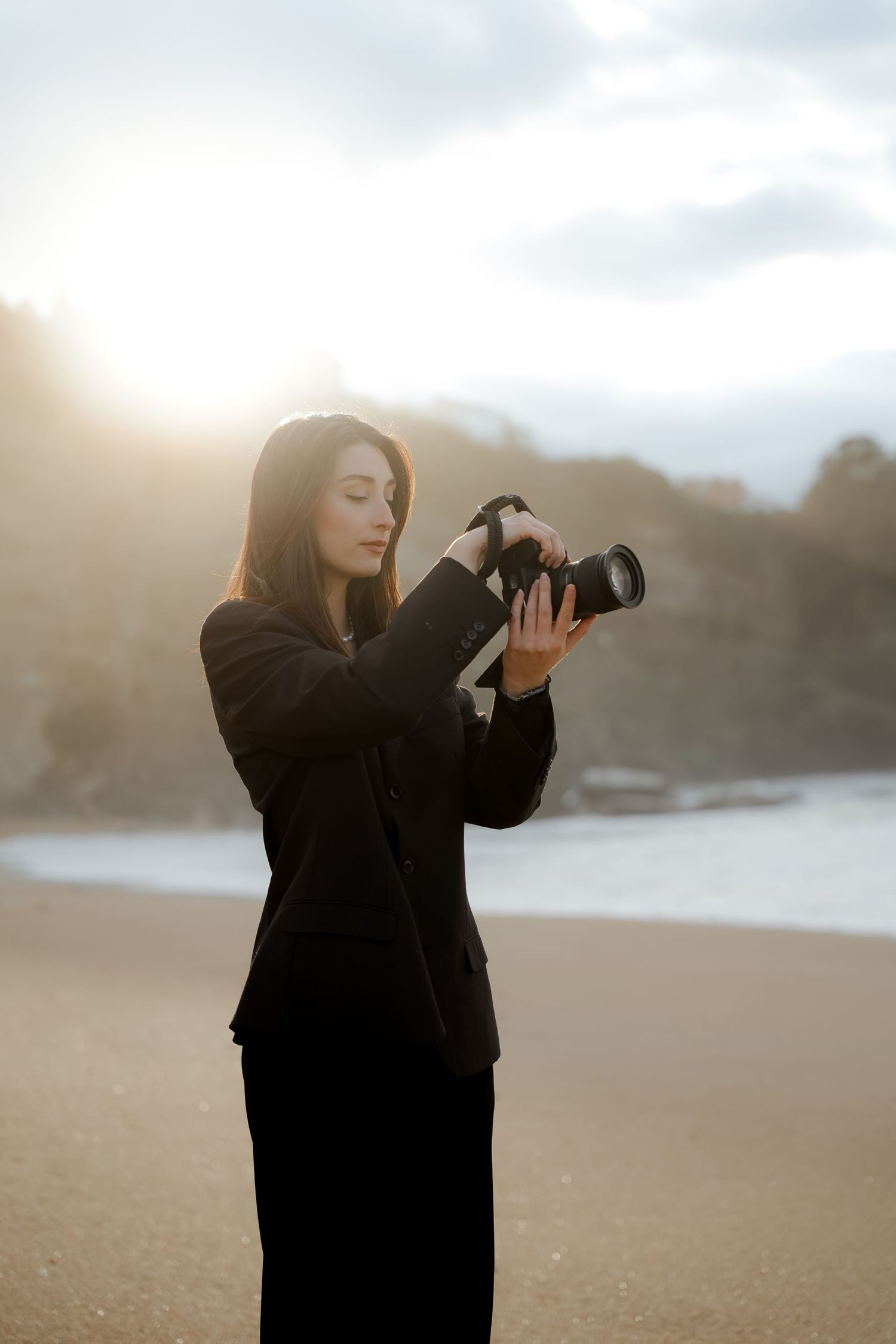 Female photographer in black suit reviewing camera on sunny beach with cliffs.