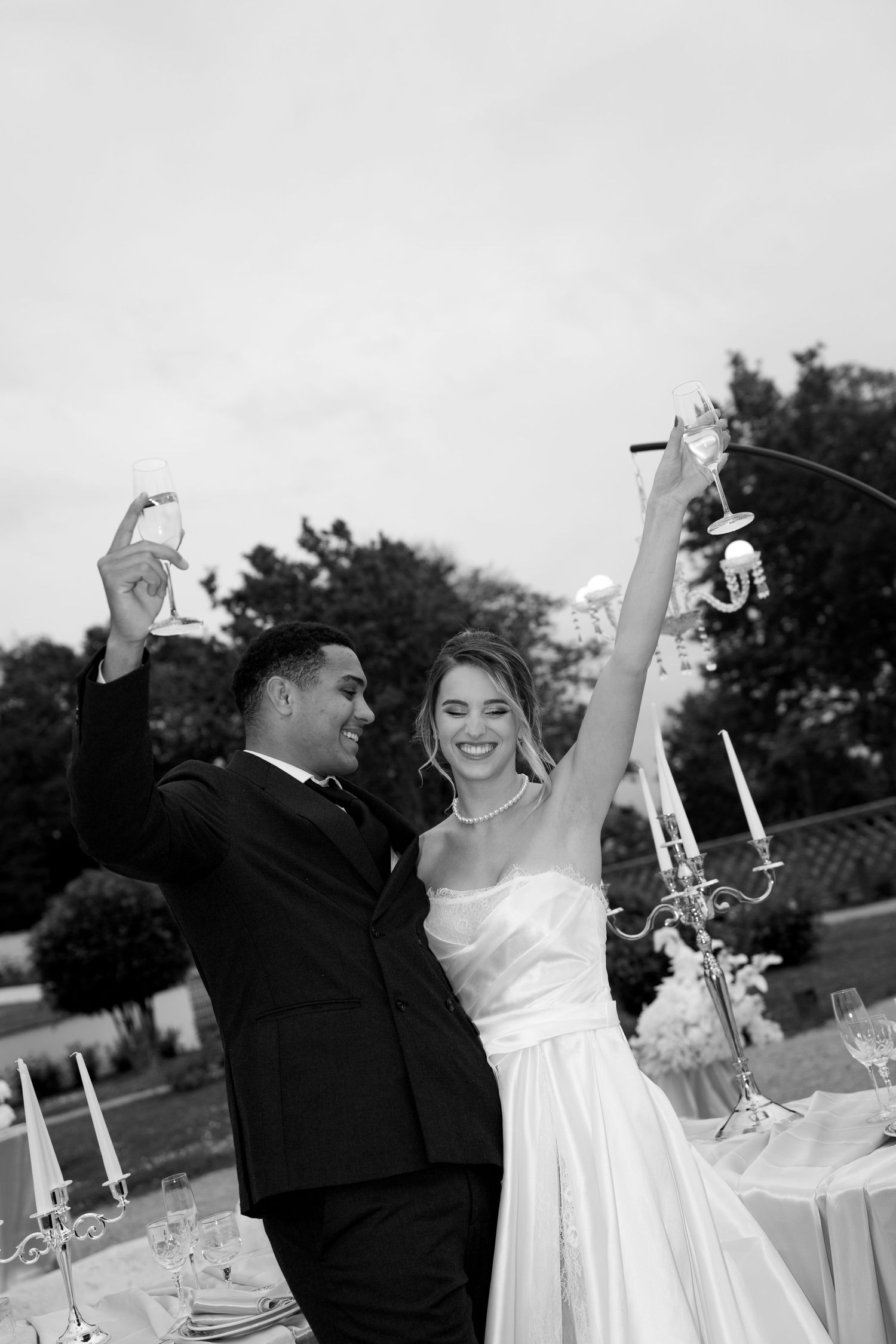 Joyful newlyweds raising champagne glasses in black and white photo at outdoor wedding reception.
