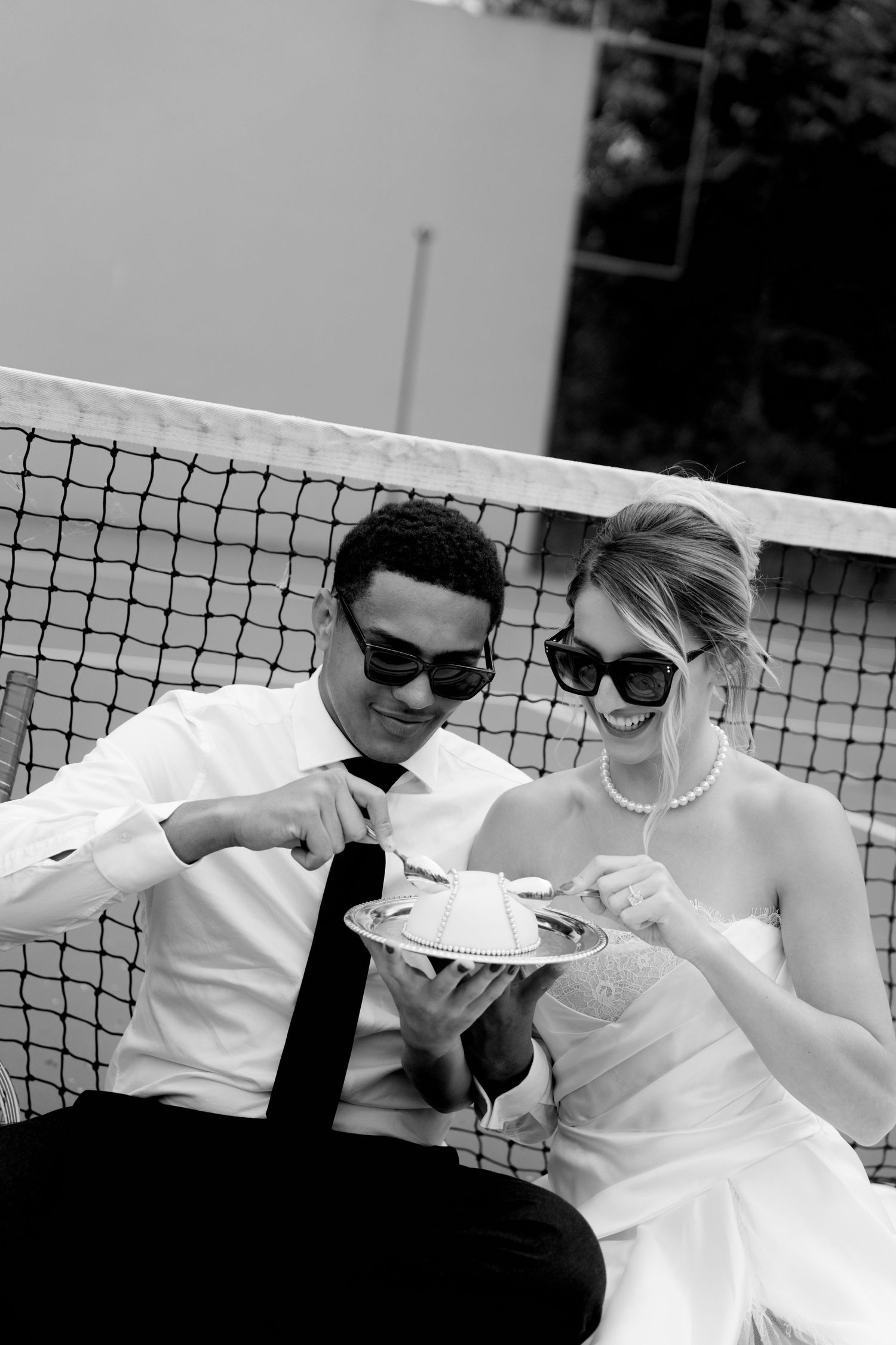 Bride and groom in sunglasses sharing wedding cake by a tennis net in a black and white photo.